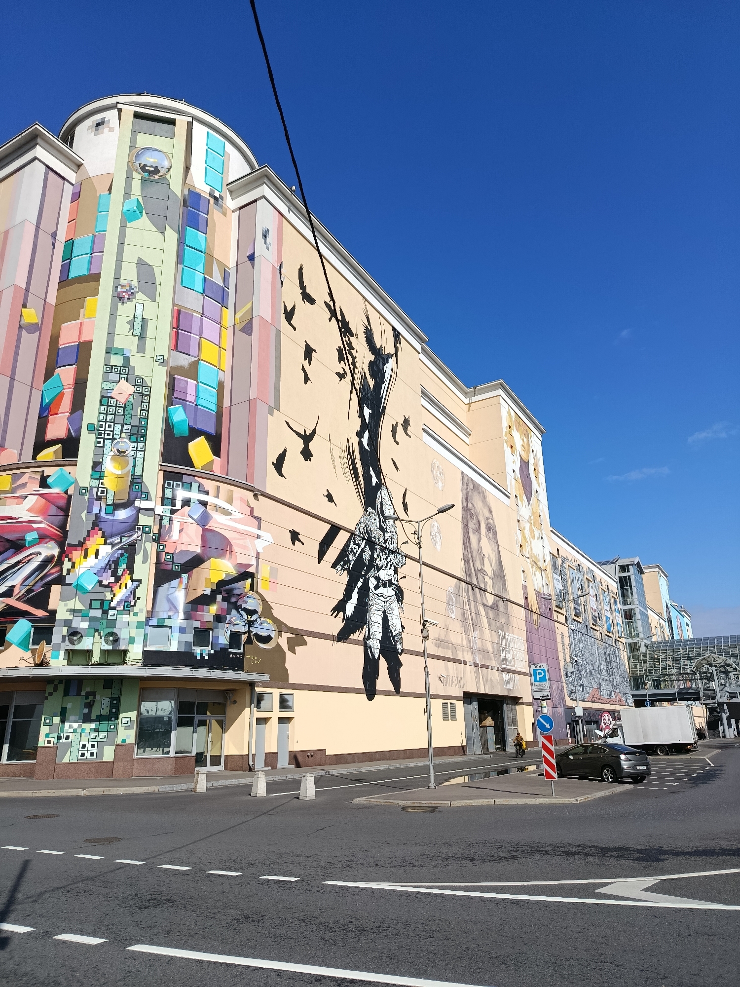 The image shows a large, colorful building in Moscow, Russia, with a vibrant mural covering its facade. The mural features abstract geometric shapes, birds in flight, and portraits of people. The building is multi-story, with windows and balconies visible. In front of the building is a street with a car parked on the side. A white truck is also visible in the background. The sky is clear and blue. The building appears to be a shopping mall or commercial complex. The architectural style is modern, with clean lines and a mix of concrete and glass. The mural is a prominent feature, adding a splash of color and artistry to the urban landscape. The street is paved and well-maintained, with traffic markings and streetlights. The overall impression is one of a lively and dynamic city.