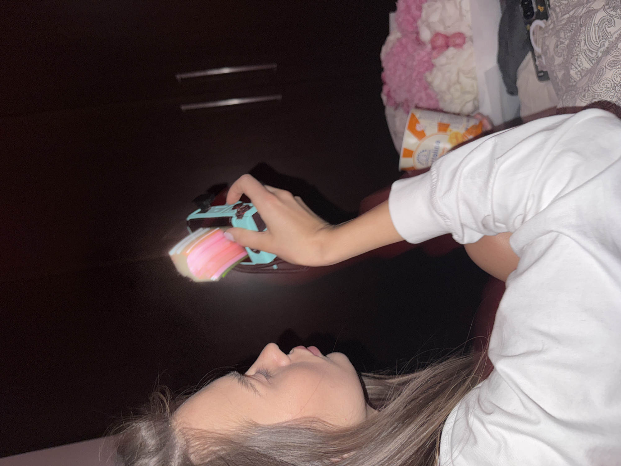 A woman lies down, holding a Nintendo Switch console. She wears a white shirt. A container of goldfish crackers is near her shoulder. A dark cabinet is behind her. A pink and white pillow is in the background. The location cannot be determined from the image.