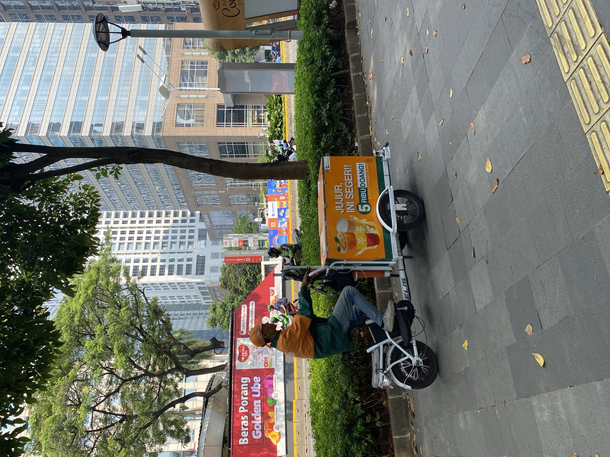 A man on a white electric tricycle is stopped on a paved sidewalk in South Jakarta, Indonesia. The man is wearing an orange and green jacket, blue jeans, and an orange cap. He is holding the handlebars of the tricycle, which has a large orange cooler attached to the back. The cooler has an advertisement for "Jujur, Ini Seger!!" and "5 Ribu Doang!". The tricycle is white with the word "ELIS" visible on the side. In the background, there are tall modern buildings, trees, and a red billboard advertising "Beras Porang Golden Ube". Other people on motorcycles are visible in the distance. The sidewalk is made of gray square tiles, and some fallen leaves are scattered on the ground.