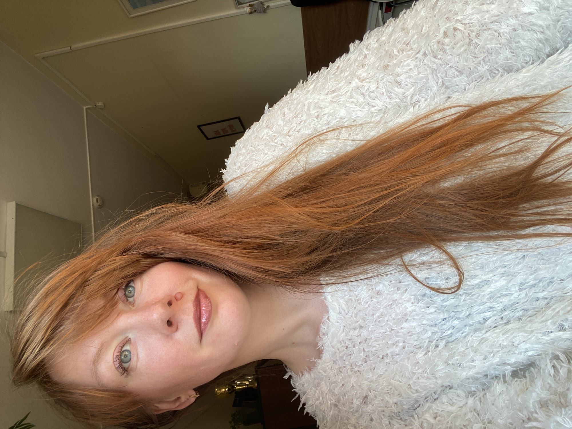 This is an indoor portrait photograph of a young woman, likely a selfie, taken in Krasnoyarsk, Russia. The woman is positioned at a slight angle, possibly lying down or holding the camera tilted, and looks directly at the viewer with a gentle smile. She has fair skin, light blue-green eyes, and long, flowing reddish-brown hair that frames her face and cascades over her shoulders. Her hair includes bangs that sweep across her forehead. She is wearing a distinctive white, highly textured, and fuzzy sweater. Visible moles are present on her face and neck.
The background shows light-colored walls and a ceiling, indicating an indoor setting. Details like a white trim along the ceiling and a small white sensor-like object are visible. A small, framed object hangs on a distant wall, but any text within it is illegible. The lighting appears soft and even, suggesting either natural daylight or well-diffused indoor illumination. The overall mood conveyed by the subject is calm and friendly.