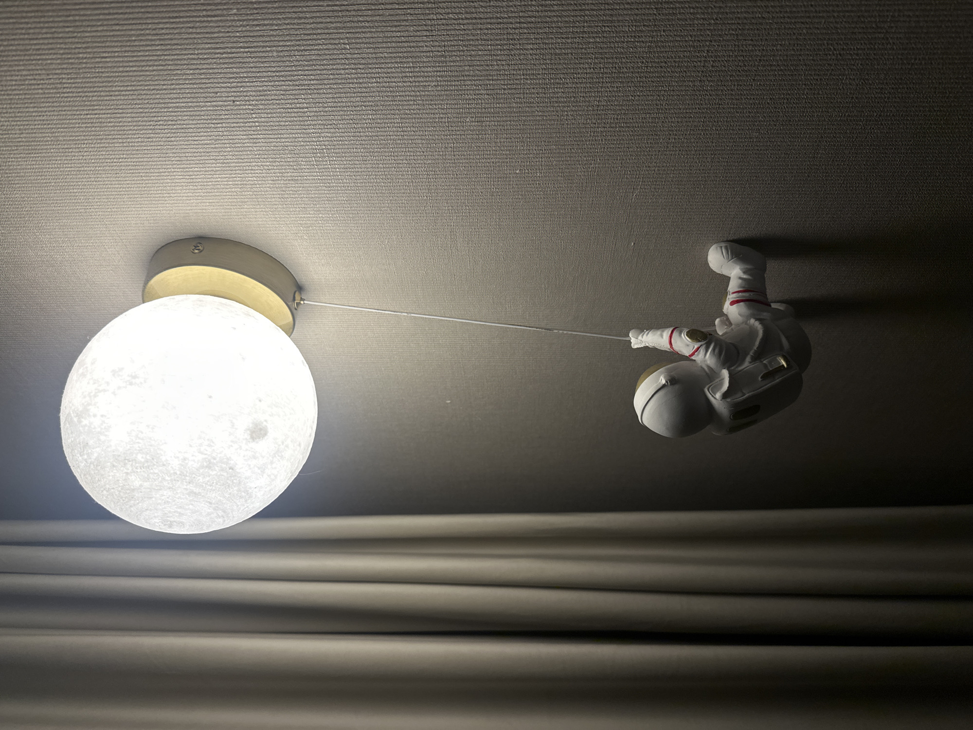 A low-angle, indoor shot depicts a lunar-shaped ceiling light emitting a bright glow on the left. An astronaut figurine is suspended in the air on the right, attached to the light fixture by a thin cable. The astronaut is wearing a white suit with red stripes and is positioned upside down. The background is a textured, light-colored ceiling and a slightly darker curtain. There's no indication of the location being Votkinsk, Russia.