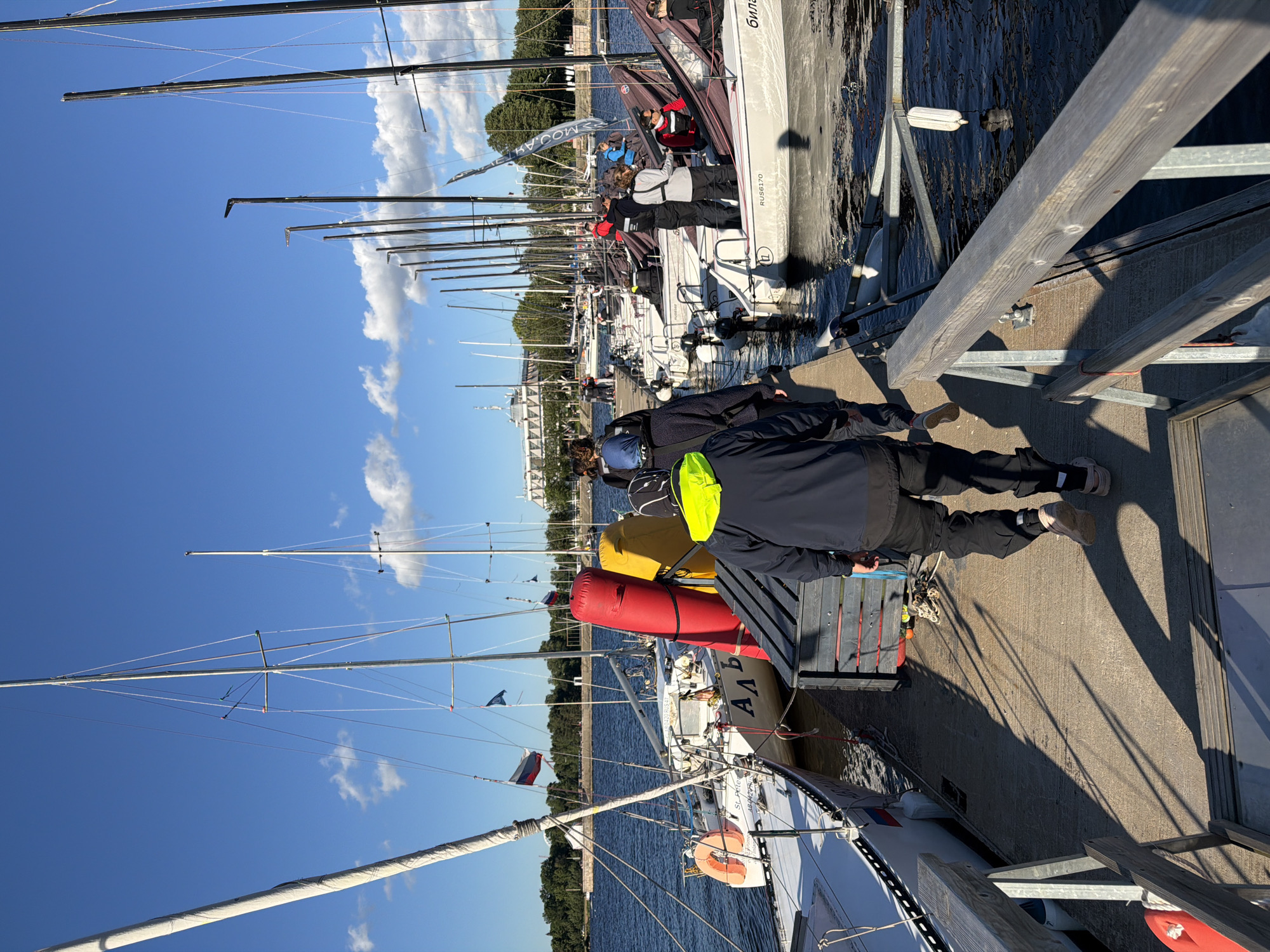 The image shows a bright, sunny day at a marina on the Neva River in Saint Petersburg, Russia. Several sailboats are docked along a pier. In the foreground, people in casual clothing and sailing gear are walking on the pier. One person in a dark jacket and black pants with a neon yellow hood is walking away from the camera. Behind them, a group of people are gathered near some of the sailboats, some of them appear to be involved in preparations or discussions related to sailing. The water of the Neva River is visible on the left side of the image. In the background, trees line the opposite bank of the river, and a large, distinctive building, possibly a museum or government building, is visible in the distance. The sky is a clear blue with a few scattered white clouds. The overall scene depicts a typical day at a marina in a coastal city, likely during a sailing event or season.