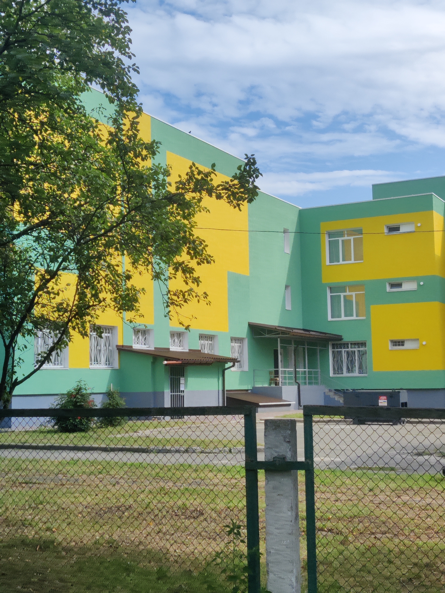 This is a photograph of a modern, two-story building with a distinctive yellow and green color scheme. The building appears to be a school or kindergarten, given its bright colors and the presence of a playground visible in the background behind a chain-link fence. The building is set against a backdrop of a blue sky with scattered white clouds. To the left of the building, a large tree with green foliage partially obscures the facade. The foreground shows a grassy area with a chain-link fence and a concrete post, suggesting the viewpoint is from across a street or public space. A few bushes are visible near the base of the building. The overall impression is that of a well-maintained, cheerful educational facility. While the exact location in Kyiv, Ukraine, is not provided in the image itself, the architectural style and the context suggest it could be in a residential or suburban area of the city. No people are visible in the image.