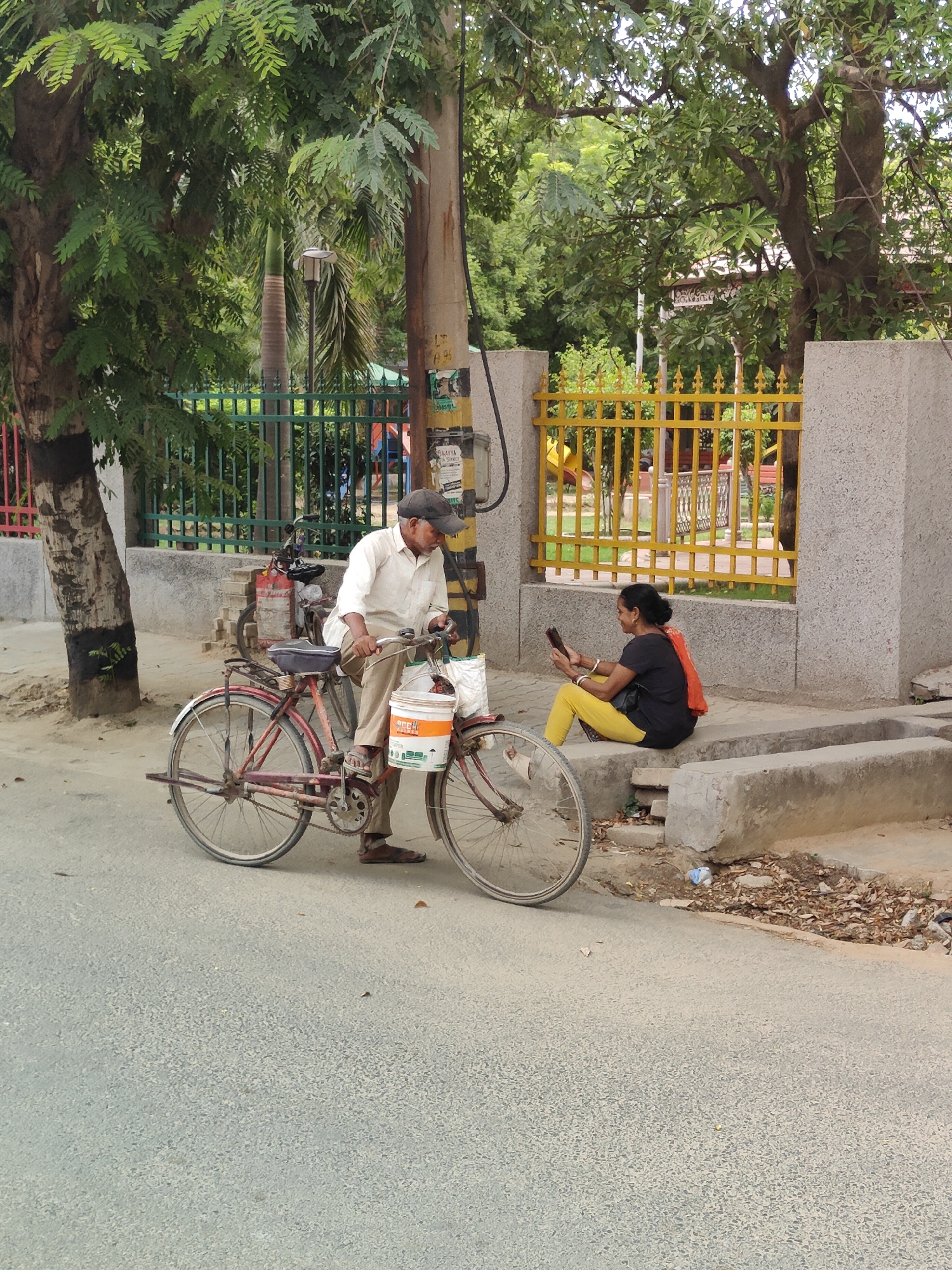The image depicts a street scene in Gurgaon, India, likely on a sunny day given the shadows. In the foreground, a man in a light-colored shirt and dark pants sits on a red bicycle. He is wearing a dark baseball cap and appears to be looking down. The bicycle has a white bucket attached to the front, and a white bag is hanging from the handlebars. The man's feet are visible, wearing brown sandals.
To the right, a woman with dark hair pulled back sits on a concrete ledge. She is wearing a black t-shirt and bright yellow pants. An orange scarf is draped over her shoulders. She is holding a mobile phone in her hands and appears to be looking at it.
Behind the man and his bicycle, another bicycle is partially visible. A green metal fence runs along the background, separating the street from what appears to be a park or playground area with yellow fencing and colorful structures. Tall trees with lush green foliage are abundant, indicating a potentially leafy urban area. A utility pole with various posters and wires stands near the man on the bicycle. The ground is paved with asphalt.
