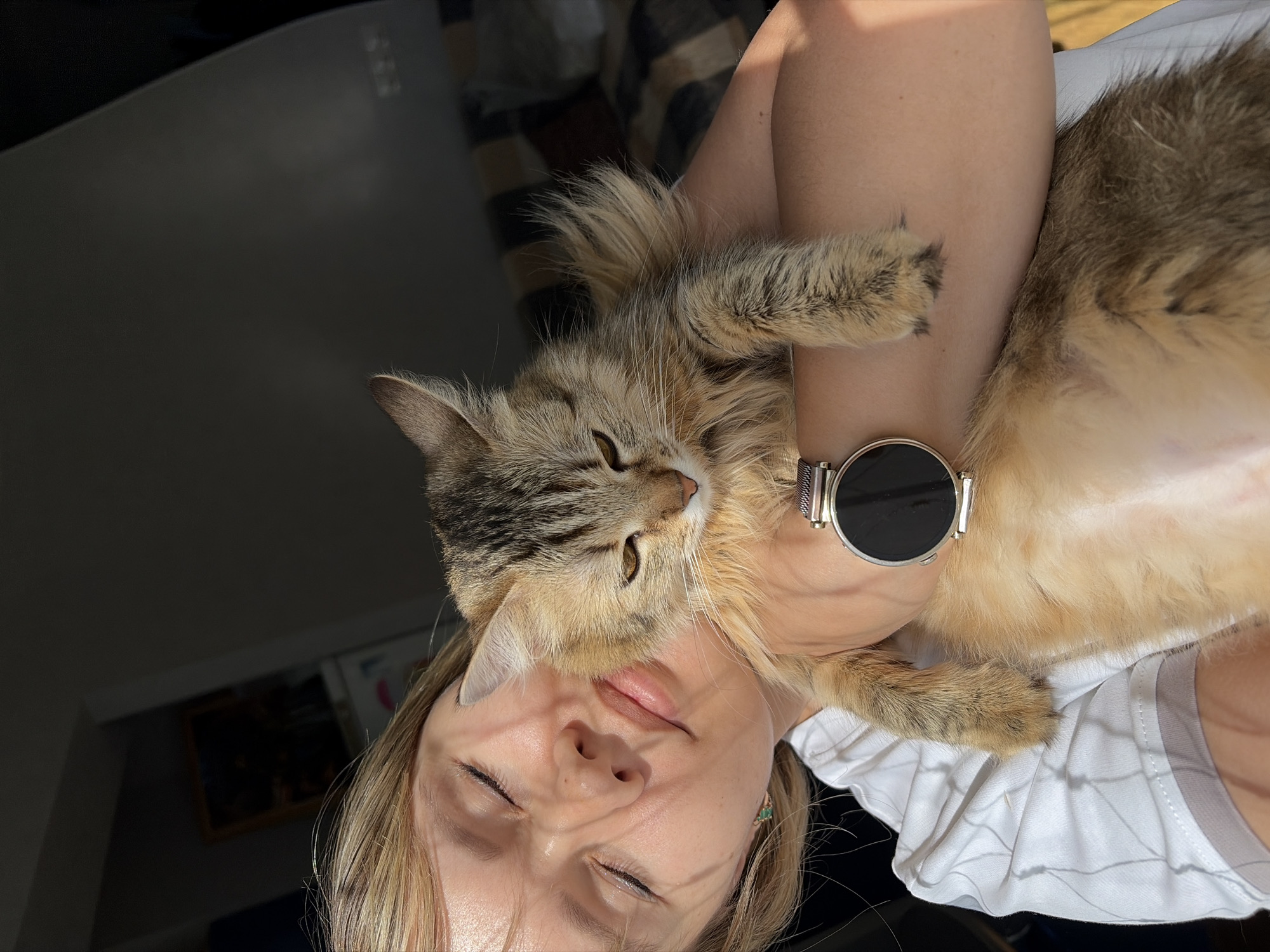A woman is holding a brown and tan tabby cat in her arms. The woman's eyes are closed and she is facing the camera. She is wearing a white shirt and a silver watch. The cat is looking to the left. The background is blurry and shows a room with a black table, a window, and a patterned blanket. The location may be in Chaikovsky, Russia.