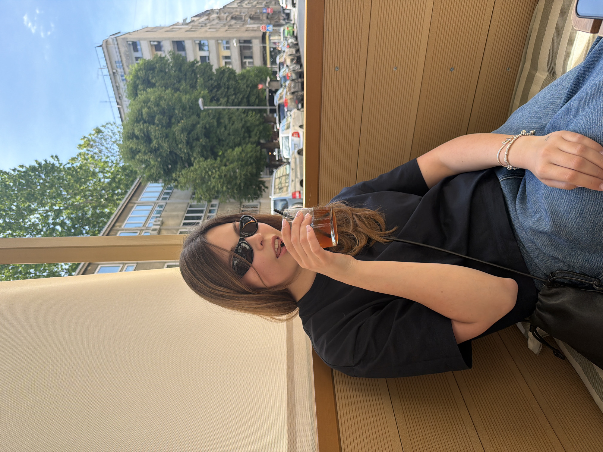 A young woman wearing sunglasses and a dark-colored top is drinking from a glass. The background includes buildings, trees, and traffic, suggesting an urban location. The seating arrangement indicates she is sitting in an outdoor cafe. The location is Belgrade, Serbia, based on architectural style.