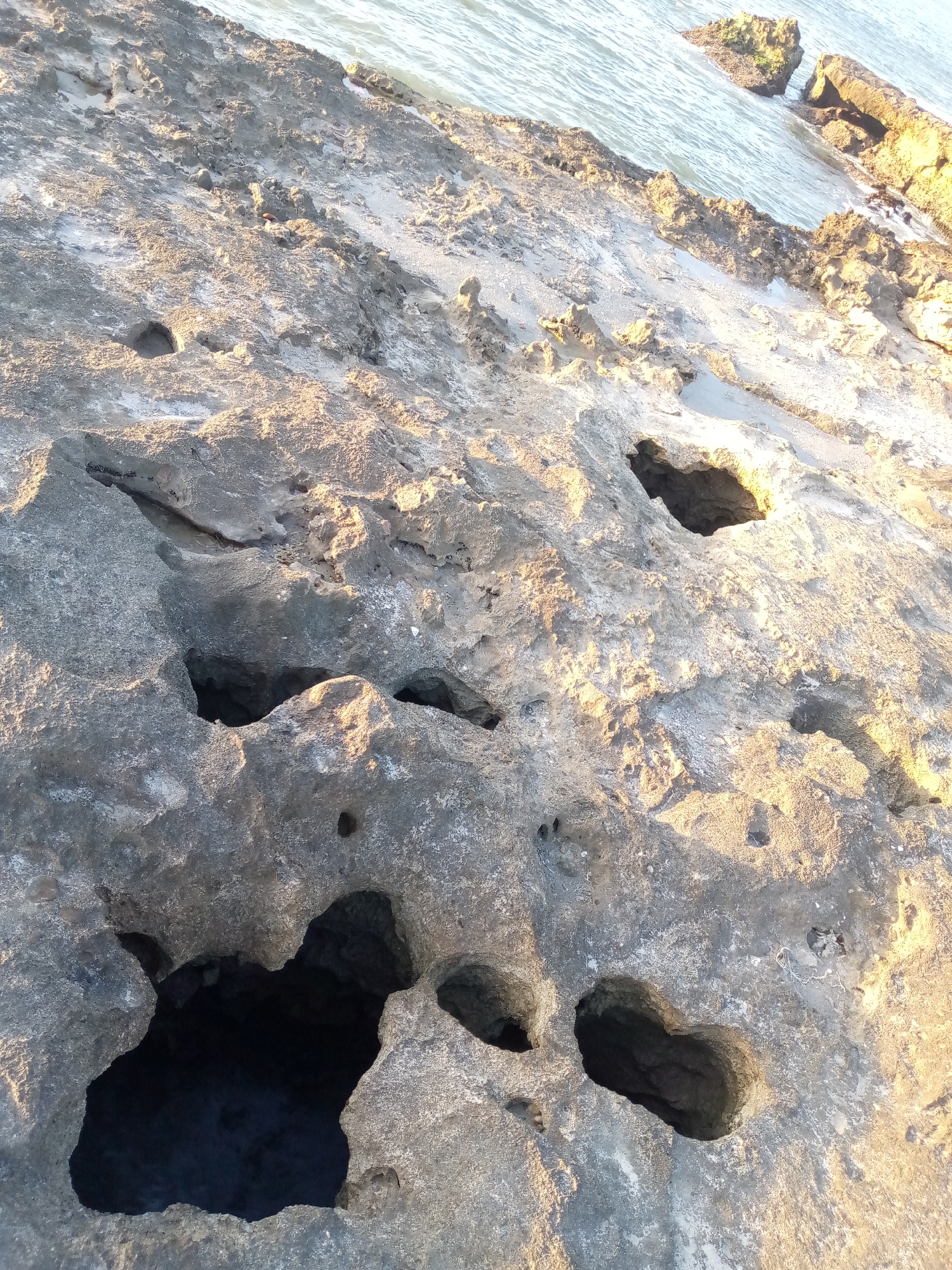 The image shows a rocky coastline with several large holes eroded into the rock. The color of the rock is mostly gray, but parts are brightly lit, likely by the sun. In the upper right portion of the image, the ocean can be seen along with more rocks. The image shows the intertidal zone of Juan Griego, Venezuela.