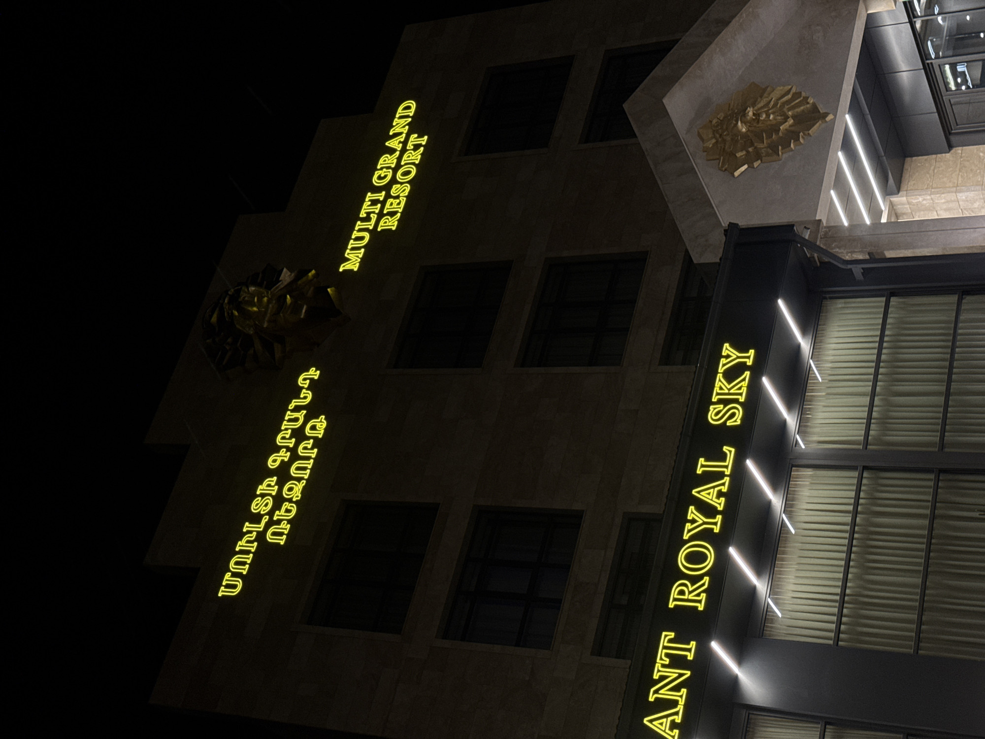 A night shot captures the exterior of the Multi Grand Resort in Tsakhkadzor, Armenia. The building's facade features the illuminated words "MULTI GRAND RESORT" and "RESTAURANT ROYAL SKY," oriented vertically. Additionally, Armenian script appears on the facade. The building's architecture is visible, including windows and architectural details. A golden artistic embellishment is above the entrance area.