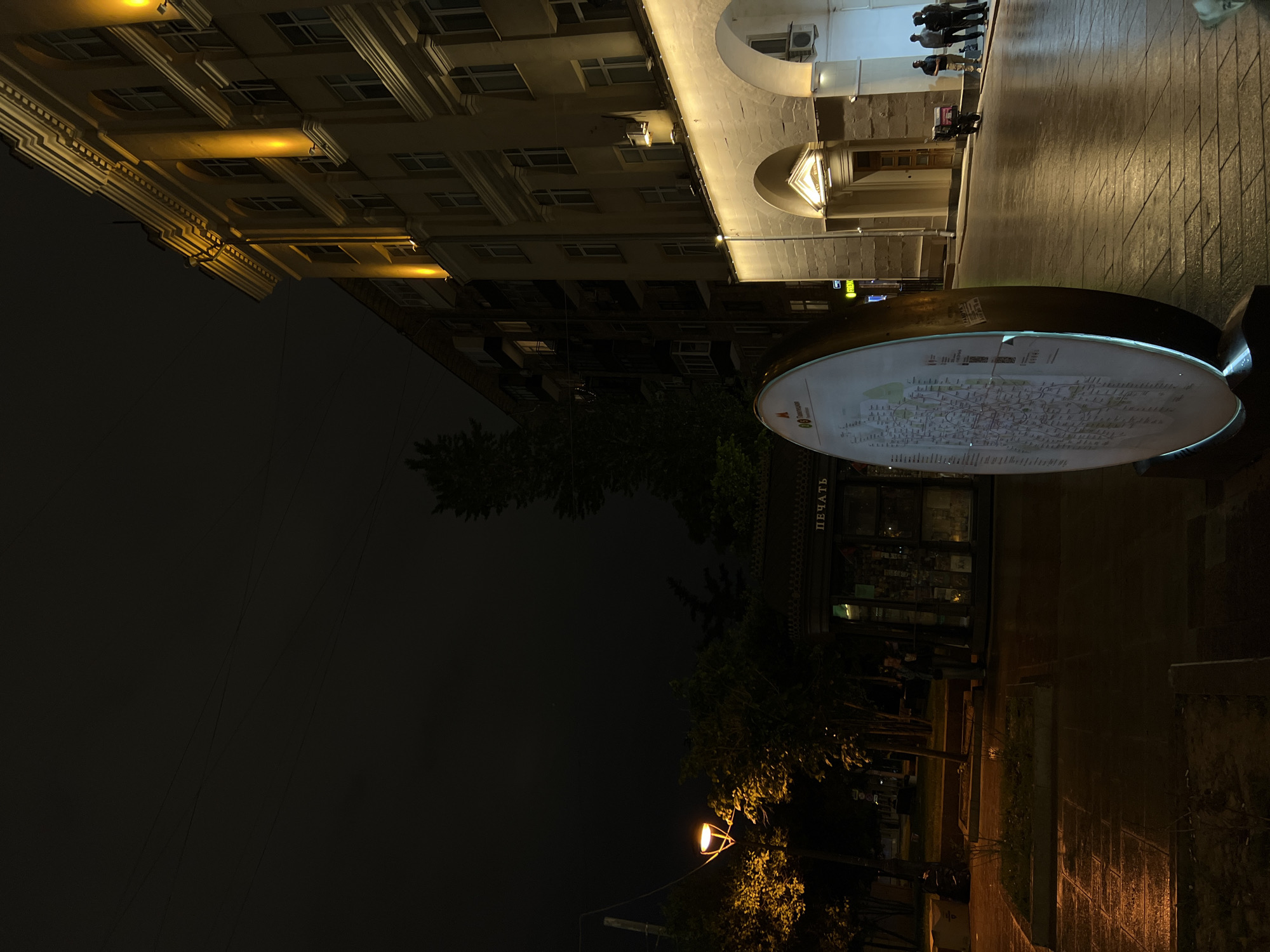 At night, the city of Moscow, Russia is captured in this image. In the upper-left quadrant, a light-colored multi-story building with rows of windows illuminated by interior lights stretches upwards. Adjacent to this building, another structure with arches and lights above a storefront extends toward the upper-right. A group of people, some in dark clothing, are walking in the same direction in the upper-right corner, while below them, the wet pavement reflects the ambient light.
In the center, a round information kiosk displays a map of Moscow. The word "ПЕЧАТЬ" (print) is legible on a storefront behind the kiosk. The bottom half of the image is characterized by a dark atmosphere with silhouettes of trees and a street lamp casting a warm glow on the surroundings. Electrical lines are visible against the dark sky. The ground level near the bottom-right is wet.