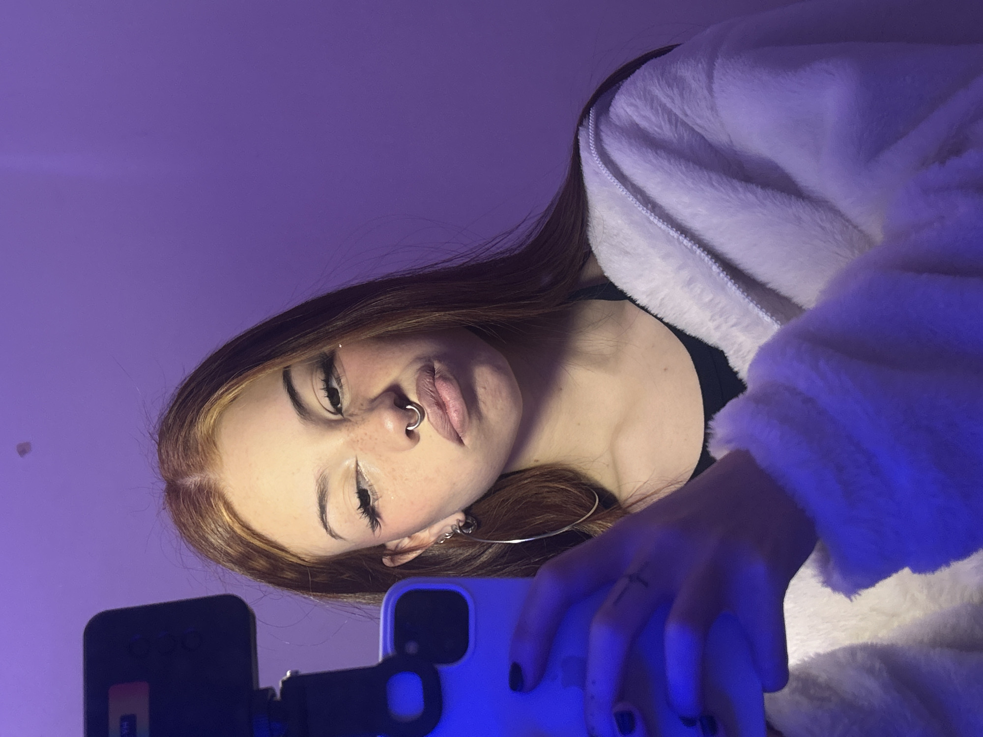 A young woman takes a selfie using a phone attached to a stand. The background is a plain purple. She is wearing a white fluffy jacket. She has auburn hair and a nose ring. There is no indication of the location being Buenos Aires, Argentina.