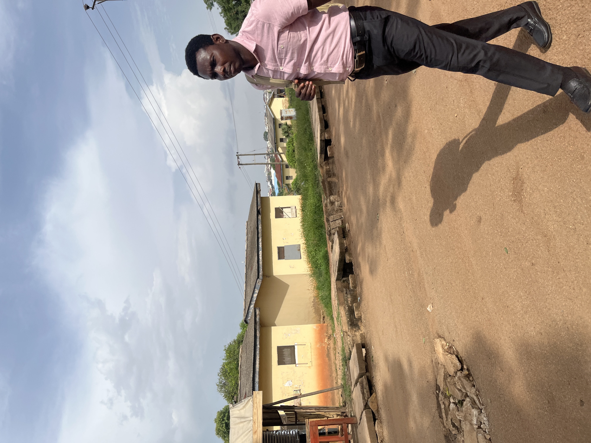 A young Black man in a light pink collared shirt, black pants, and black loafers walks on a dirt road in Rafin Par, Nigeria. He is carrying a light-colored shoulder bag. In the background, there are several yellow buildings with corrugated metal roofs and a cloudy sky. Some vegetation is visible between the buildings. The road appears unpaved and has patches of broken concrete.