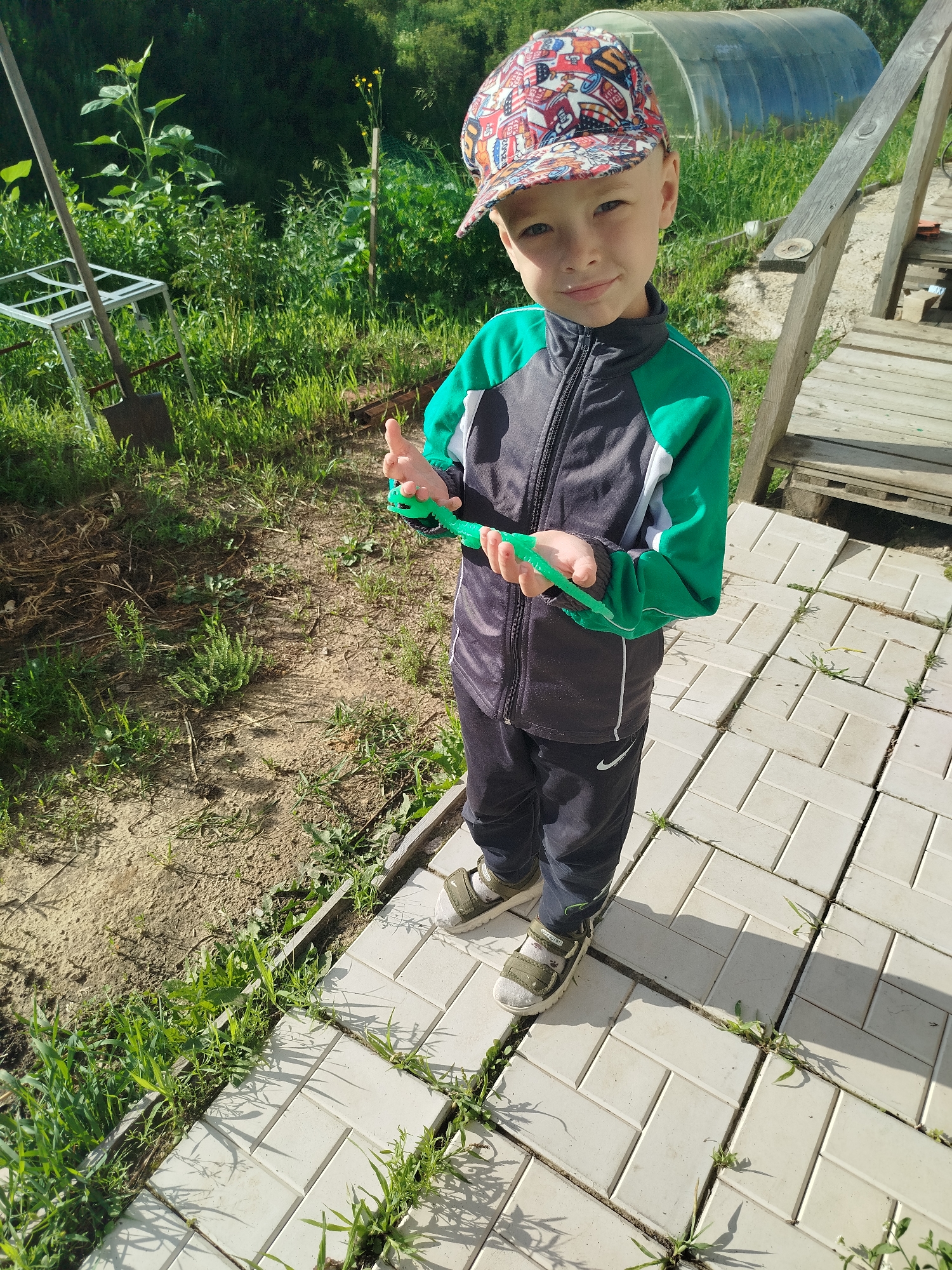 A young boy wearing a colorful baseball cap, a green and gray zip-up jacket, and dark pants stands on a light-colored tiled patio. He is holding a green, segmented toy in his hands. The background consists of lush green vegetation, a wooden structure with railings, and a greenhouse. The scene appears to be outdoors in a rural or suburban setting. The specific location in Zavodoukovsk, Russia, cannot be determined from the visual information alone.