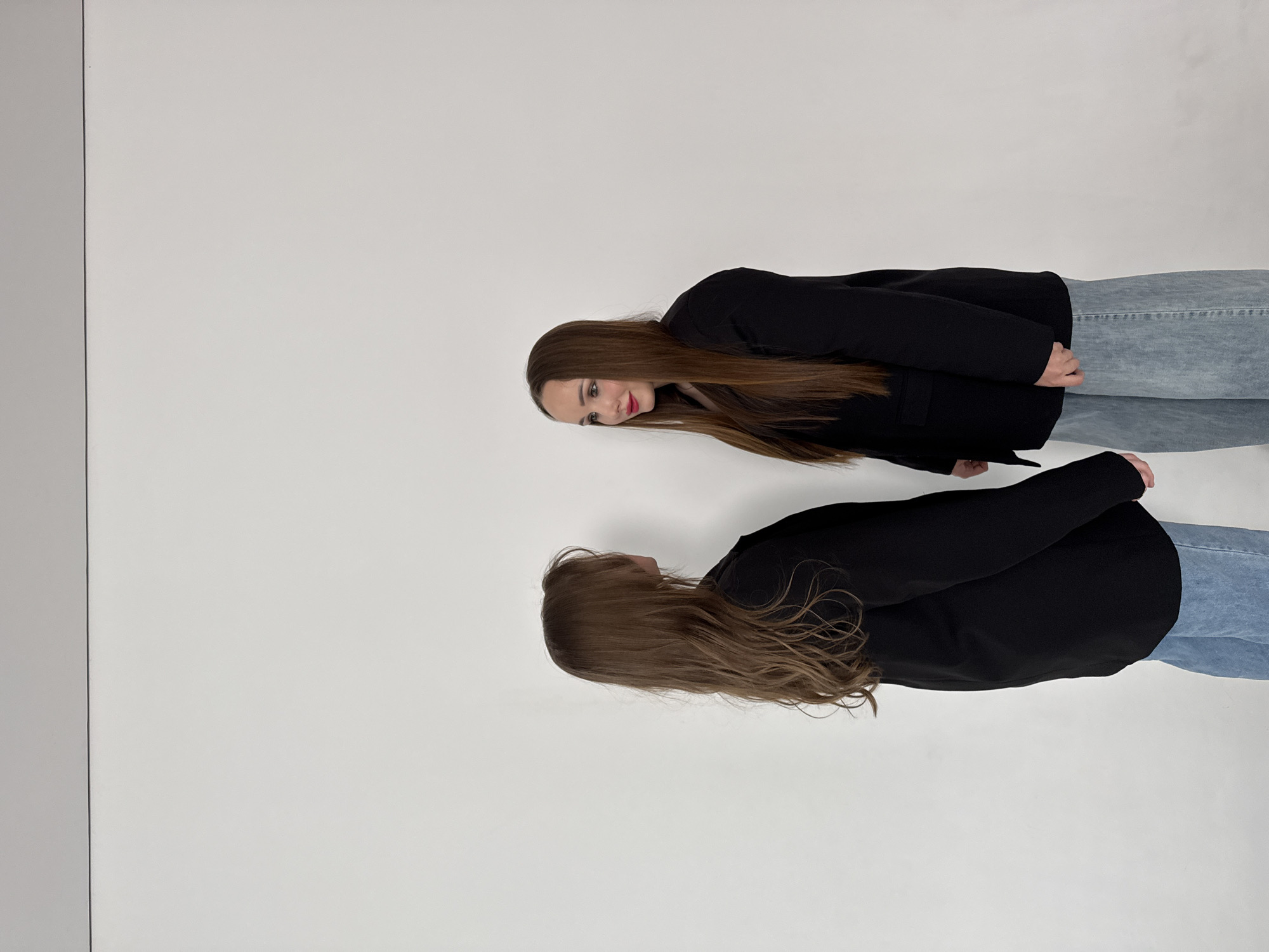 A woman in a black blazer and light blue jeans stands in front of a white background, facing her reflection on a gray surface that mimics a mirror on the left side of the frame. Her hair is long, straight, and brown. The location is a studio or photo shoot setting. There is no indication that this is in the city of Chaikovsky, Russia.