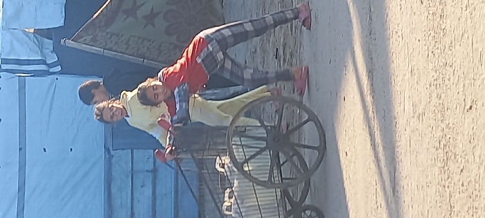 In Khan Yunis, Palestinian Territory, three individuals, appearing to be children or young adolescents, are observed moving a handcart on an unpaved, light-colored dirt surface under natural daylight. A girl wearing a red long-sleeved shirt and gray-and-white plaid pants is pushing a metal-framed, two-wheeled cart from the front right, leaning into it with both hands. The cart contains light-colored bundled items. Behind her, another girl in a yellow long-sleeved top and pants, wearing glasses, holds the cart's handles, also participating in pushing its movement. A third individual, wearing dark clothing, stands behind the girl in yellow. In the background, a large light blue or white tent structure is visible, along with a patterned green blanket displaying star motifs, hanging on a vertical support. Shadows are cast on the ground from sunlight.