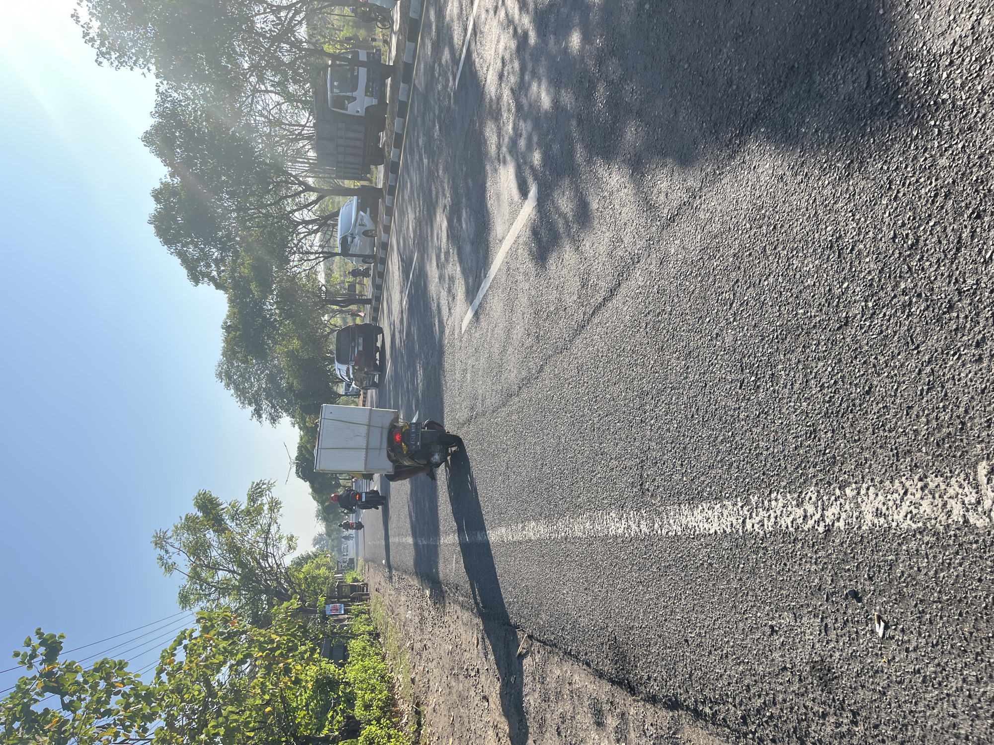 The image shows a road in Siring, Indonesia, under a clear blue sky with bright sunlight casting shadows. A motorcycle carrying a large white box is in the foreground, traveling on the asphalt road. There is a white line painted down the center of the road. To the left of the road is a dirt shoulder with some vegetation and a few trees. To the right of the road, there are more trees lining the side, and several vehicles, including cars and a truck, are visible. In the distance, more trees and buildings can be seen. The overall scene depicts a typical street in a tropical or subtropical urban or semi-urban environment.