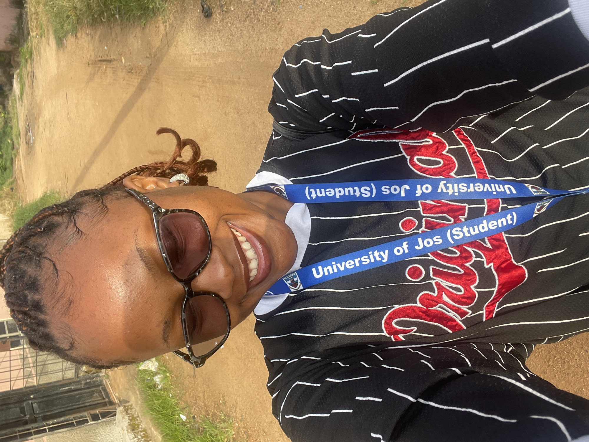 A young Black woman with braided hair and sunglasses smiles as she takes a selfie outdoors. She is wearing a black and white striped jersey with red lettering and a blue lanyard around her neck that reads "University of Jos (Student)". The background is a dirt road lined with dry grass, and a building is visible to the left. The scene appears to be in Rafin Par, Nigeria.