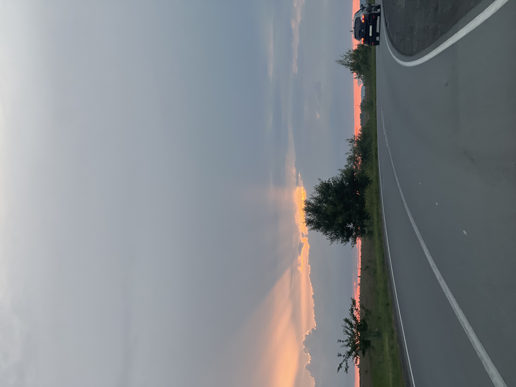 The image shows a road with a car on it, on the side of a grassy field, under a cloudy sky. The sky is dark grey with a band of pink and orange light on the horizon, suggesting it is either sunrise or sunset. The road is grey asphalt with a white line marking the lane. A black car is visible on the right side of the road, moving away from the viewer. There are trees and bushes on the left side of the road and in the distance. The overall scene appears calm and serene. There is no visible text in the image.
