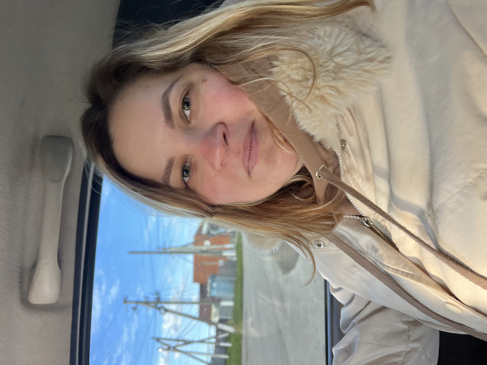 A woman with blonde hair and green eyes is taking a selfie in a car. She is wearing a beige jacket with a faux fur collar. Outside the window, utility poles and a building are visible, suggesting a residential area in Novopokrovskaya, Russia. The sky is blue with scattered clouds. A white handle is visible inside the car.