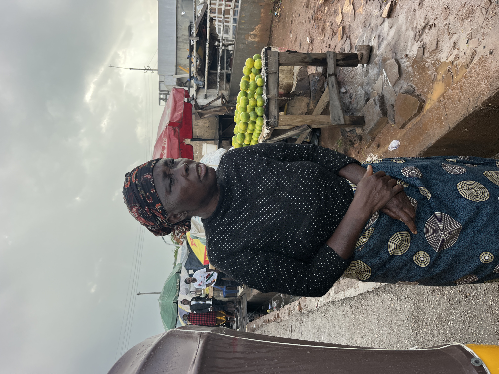 An outdoor medium shot captures a Black woman standing in a market setting. She is looking to her left and has her hands clasped in front of her. She wears a black top with white polka dots and a patterned skirt. Her head is wrapped in a colorful headscarf. To her right, a wooden table is piled high with green citrus fruits, likely limes. In the background, other market stalls and people are visible, including umbrellas and awnings. The ground appears wet and muddy, suggesting recent rain. A brown and yellow vehicle is partially visible on the left side of the frame. The sky is overcast and gray.