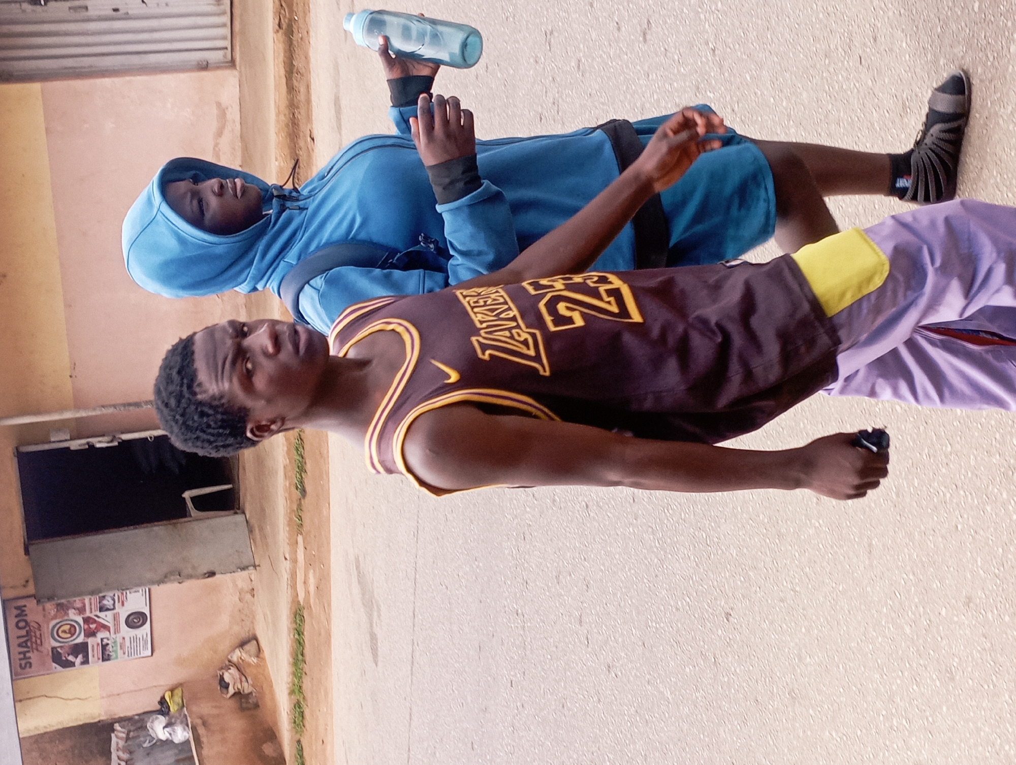 This image depicts a street scene in Jos, Nigeria. In the foreground, a young man walks towards the viewer's right. He is wearing a dark purple sleeveless basketball jersey with yellow trim and the number "23" printed in yellow on the front, along with the word "LAKERS" in yellow. He also wears light purple pants with a yellow patch on the left side and black socks with grey sandals. His left hand is slightly outstretched. Behind him and to his right, a young woman walks away from the viewer. She is wearing a bright blue hooded jacket with black trim, and dark blue shorts. She carries a clear blue water bottle in her right hand, which is held near her hip. A dark backpack is visible on her back. The background shows a building with a light pink facade and a darker, possibly brown, trim. A sign with "SHALOM FEED" is visible on the building. An open doorway reveals a dark interior. The ground is paved with asphalt.