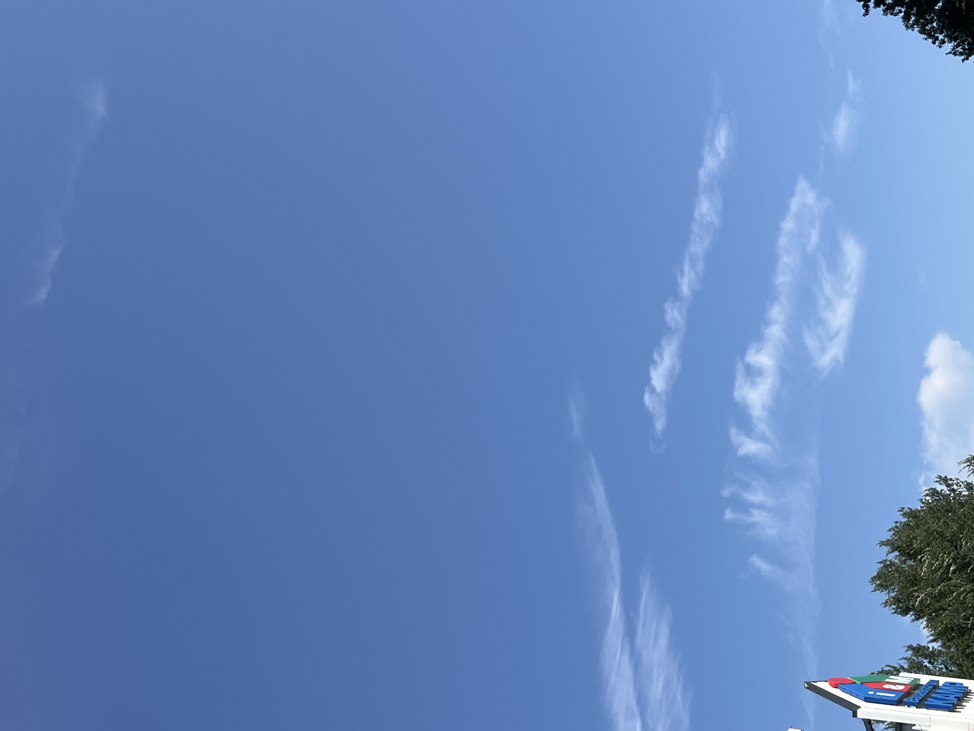 A low-angle shot captures a bright blue sky with scattered wispy clouds. In the lower left corner, the top portion of a white sign with colorful geometric shapes and text is visible. The text on the sign appears to be in a blue font with a white outline, and reads "i & M" followed by other text below. To the right of the sign, the dark green foliage of trees reaches into the frame. The overall scene suggests an outdoor environment, possibly a commercial area given the sign. The time of day appears to be daytime, with the sun illuminating the sky.