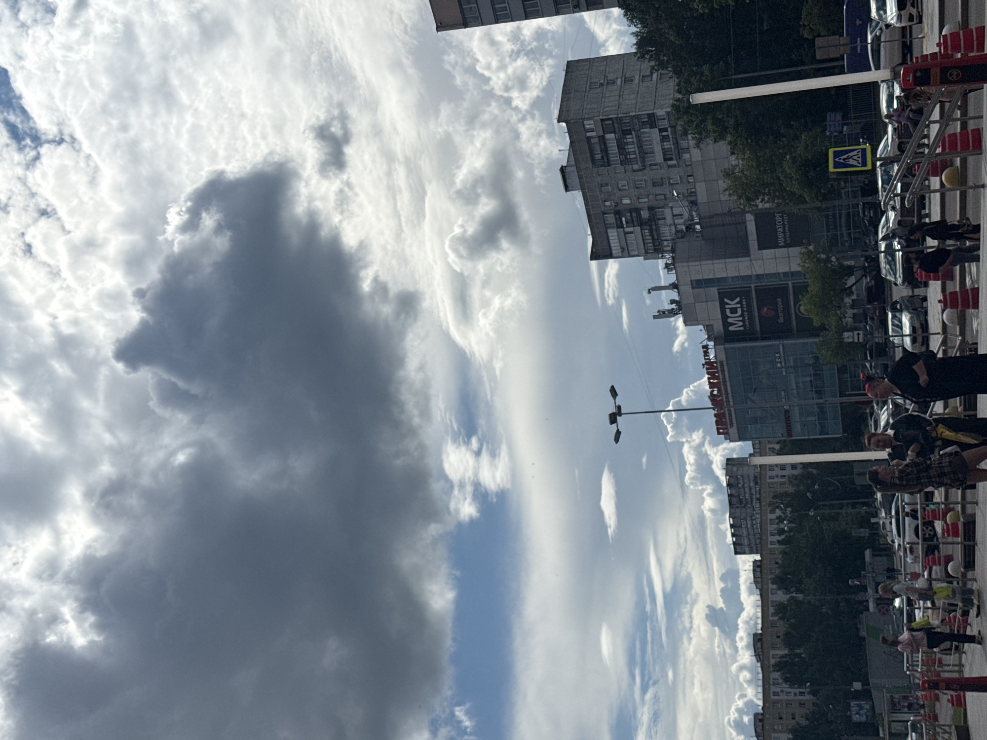 A street scene in Moscow, Russia, is captured from a low angle looking upwards towards a dramatic sky filled with a mix of dark, ominous storm clouds and bright, wispy cirrus clouds. Sunlight breaks through in places, illuminating the fluffy textures of the clouds.
In the foreground, a sidewalk is bordered by red and white safety barriers, suggesting ongoing construction or event setup. A yellow pedestrian crossing sign is visible on a pole. Various cars are parked along the street, some partially obscured by the barriers.
Several people are present in the scene, some walking along the sidewalk, others appearing to be observing or interacting. They are dressed in casual, warm-weather clothing. The overall atmosphere appears to be a bright day with the possibility of an approaching storm.
Buildings line the street, featuring a mix of architectural styles, with some appearing to be apartment blocks and others modern commercial structures. Visible text includes "MCK LOUNGE-BAR" and what appears to be Russian Cyrillic script on the buildings, such as "МИРАТОРГ" (Miratorg) and "Яйцотория" (Yaytcotoriya). A street lamp with multiple lights is also visible against the sky. The time of day appears to be late afternoon or early evening, given the angle of the light and the dramatic cloud formations.