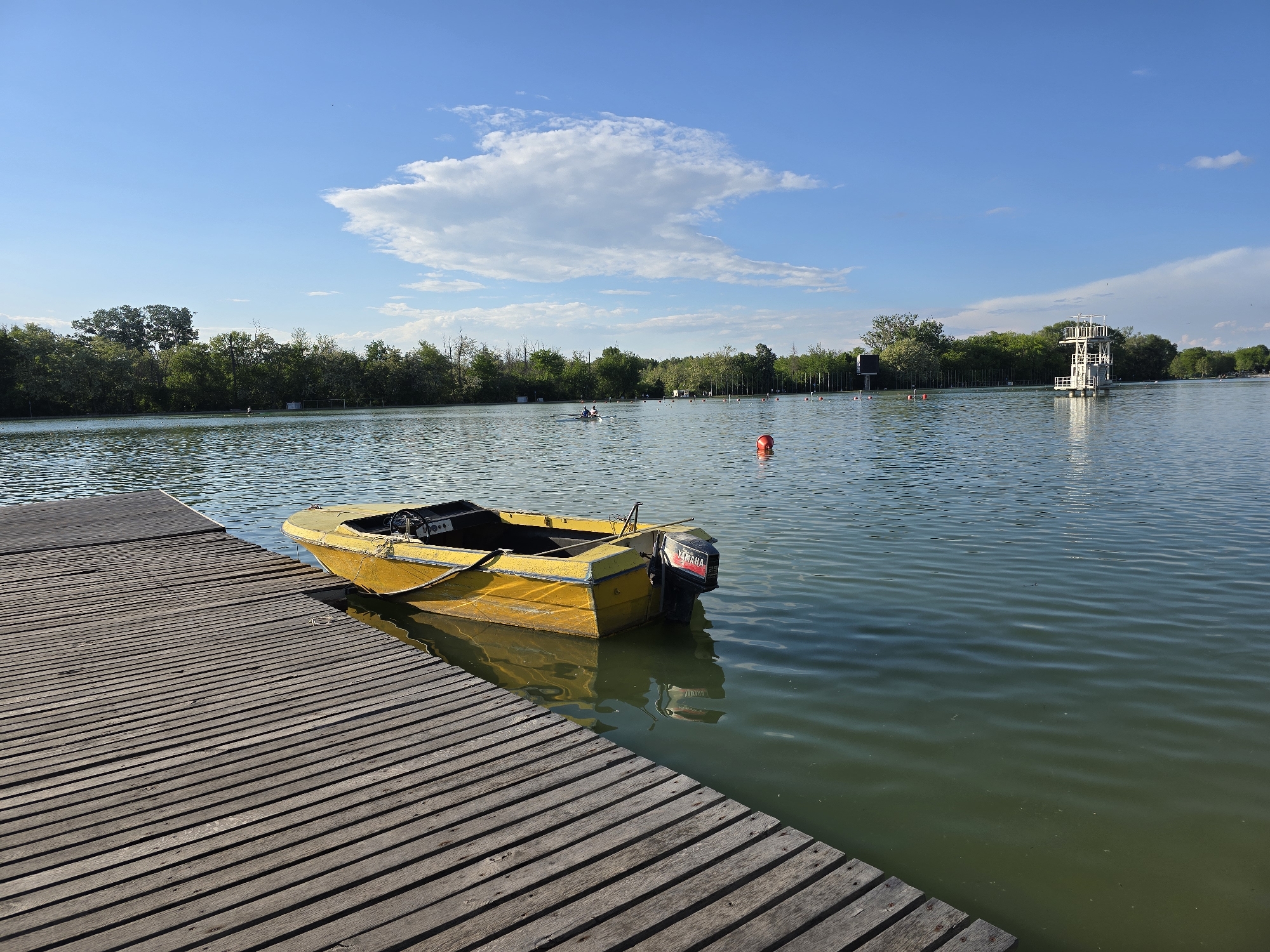 The image depicts a yellow motorboat docked on a wooden pier. The pier is made of wooden planks running horizontally across the bottom left of the image. The boat is tied to the pier with a rope. A Yamaha outboard motor is attached to the back of the boat. The water surrounding the boat is calm, with reflections of the boat and sky visible. In the background, a lake stretches to the horizon, with trees lining the far shore. A white tower structure is visible on the right side of the background. The sky is blue with a few scattered clouds. The image was taken in the city of Unknown, Unknown.