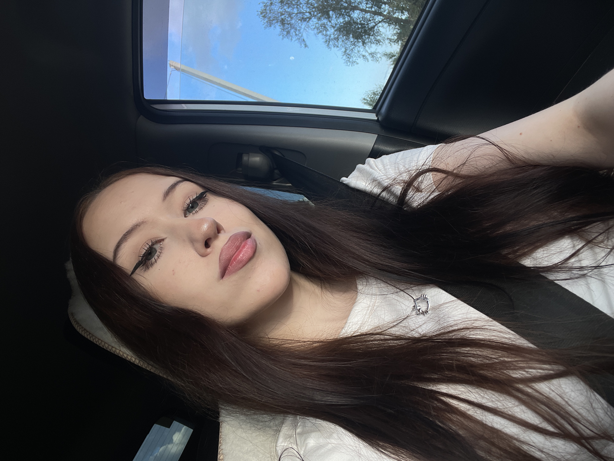 A young woman with long, dark hair is taking a selfie in a car. She is wearing a white t-shirt and a silver pendant necklace. The sunroof of the car is open, revealing a blue sky with some clouds and trees in the distance. The scene appears to be set during the daytime. The woman's makeup is visible, with winged eyeliner and light pink lipstick. Her expression is relaxed. The car's interior, including a seatbelt, is also visible. The location is identified as Gavrilovka, Russia.