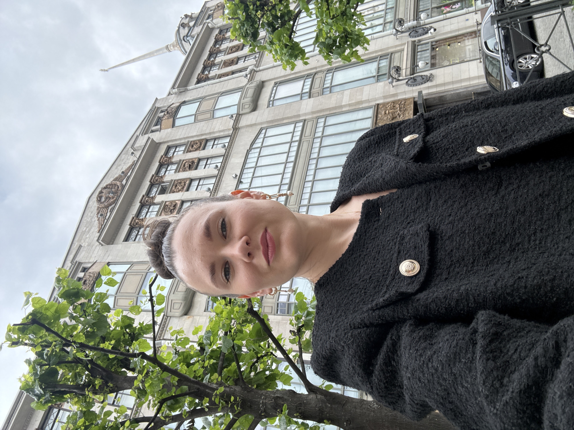 A woman is taking a selfie in front of a grand building in Saint Petersburg, Russia. The building, which appears to be a department store or a historic landmark, has a light-colored facade with numerous windows and ornate architectural details. A tall, slender spire is visible on the right side of the building. A leafy tree is in the foreground on the left, partially obscuring the woman and the building. The woman is wearing a black textured jacket with gold buttons and pearl earrings. Her hair is pulled up in a bun. A black car is parked on the street in front of the building. The sky is overcast.