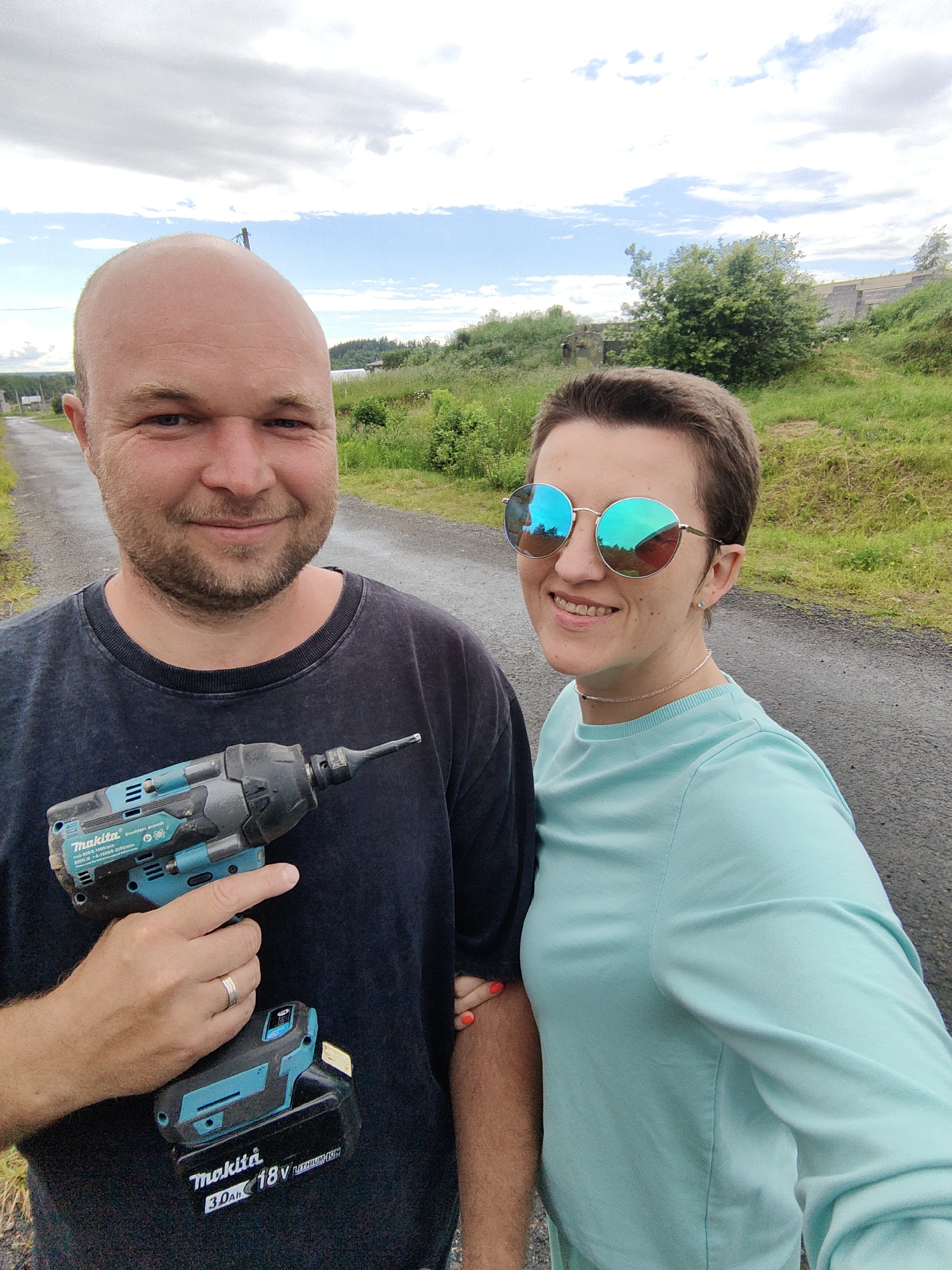Two people, a man and a woman, are standing side-by-side on a gravel road. The man, on the left, is bald with a beard and is wearing a dark t-shirt. He is holding a blue and black Makita impact driver with a battery attached. The woman, on the right, has short brown hair and is wearing a light blue long-sleeved shirt and sunglasses with blue reflective lenses. They are both smiling. The background is a rural landscape with green grass, trees, and a cloudy sky. The location is identified as Гаранскі сельскі Савет, Belarus.