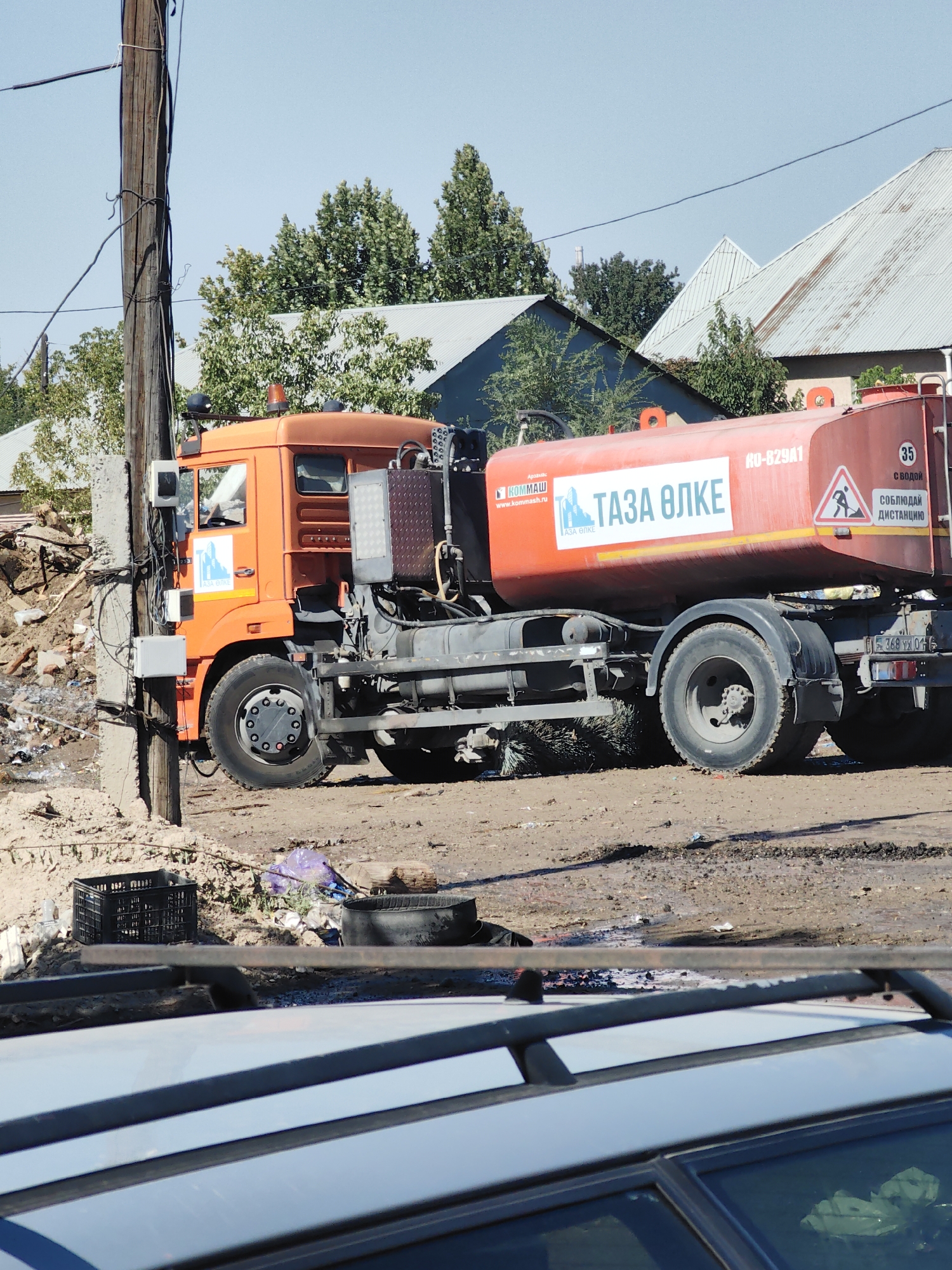 An orange tanker truck with a red tank is parked on a dirt road in Shymkent, Kazakhstan. The truck has the logo "TAZA ELKE" on its side, which translates to "Clean Land" in Kazakh. The truck is surrounded by debris, including bricks, wood, and trash. In the background, there are houses with metal roofs and trees. The sky is clear and blue. A wooden utility pole with wires is to the left of the truck. A portion of a silver car is visible in the foreground, obscuring the lower part of the scene. There is no visible human activity or interaction in the image.