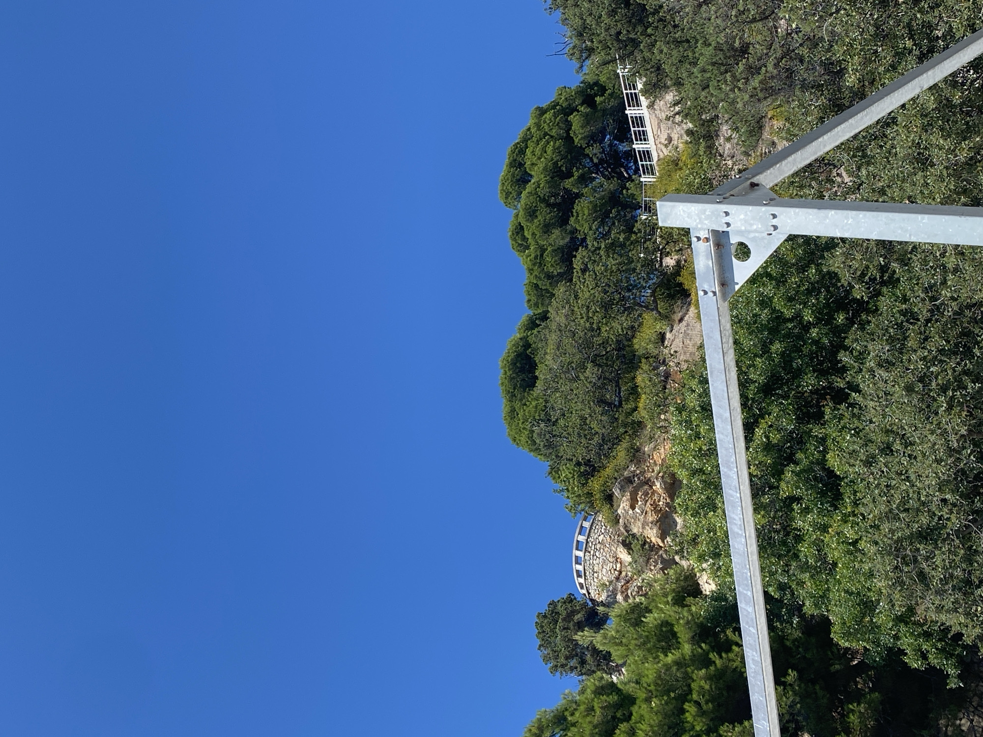 The image shows a clear blue sky above a rocky, tree-covered hillside. On the top of the hill, there is a circular stone structure with a white railing. A white railing also runs along a path on the right side of the hill. In the foreground, a portion of a metallic structure, possibly part of a larger installation, is visible, with its support beams and horizontal bars made of galvanized metal. Lush green trees and bushes cover the lower and middle sections of the hillside. The location is identified as Гаспринский поселковый совет, Ukraine.