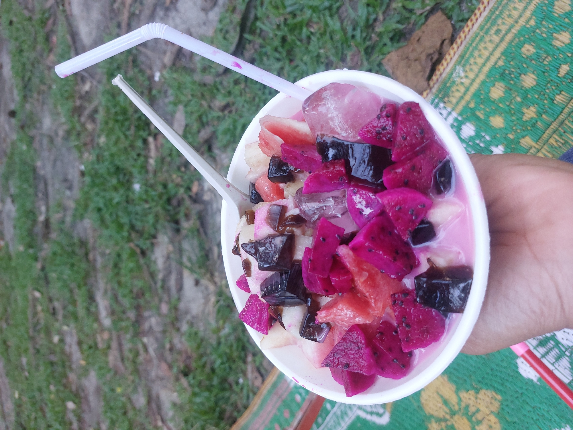 A person holds a white bowl filled with a fruit salad in the City of Pematang Siantar, Indonesia. The salad contains chunks of dragon fruit, watermelon, cubes of dark jelly, and ice. Two straws, one white and one pink, are inserted into the bowl. The person is seated on a green woven mat outdoors, with grass and dirt visible in the background.