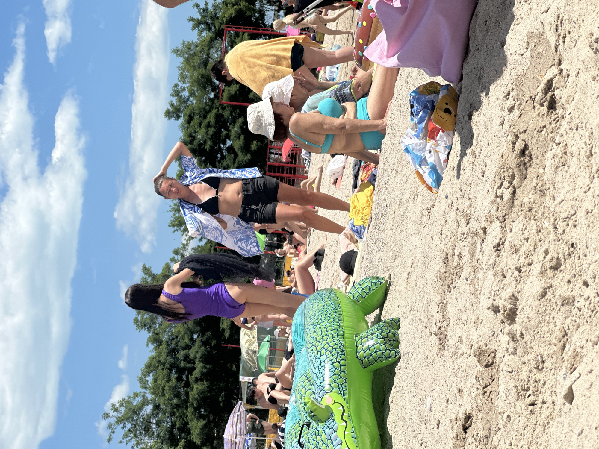 The image depicts a sunny day on a sandy beach, likely in Odesa, Ukraine, given the context. Numerous people are scattered across the beach, engaged in various activities. In the foreground, a large, inflatable green alligator is partially visible. Several beachgoers are dressed in swimwear, with some relaxing on the sand, others standing or walking. Umbrellas and towels are strewn around, indicating typical beach leisure. The environment includes a clear blue sky with scattered clouds and lush green trees in the background, suggesting a warm climate. The overall scene conveys a sense of a crowded public beach during peak season.