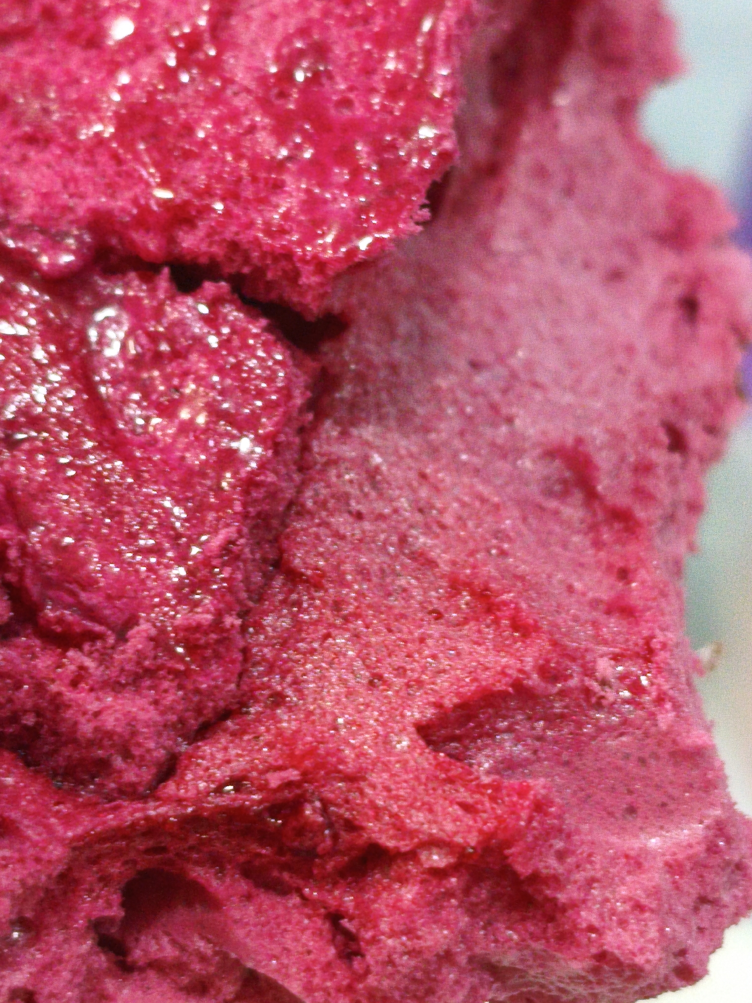 The image displays a close-up, macro view of a brightly colored, textured substance, likely a food item such as sorbet, ice cream, or a sponge cake, with a rich magenta or deep pink hue. The surface is uneven and porous, with visible air bubbles and glistening droplets, suggesting a cold or moist texture. The lighting highlights the shiny surfaces, creating bright specular reflections. The background is out of focus and appears to be a muted light color, possibly a pale blue or white. There is no indication of any specific location within Orenburg, Russia, or any elements that would suggest an urban environment. The image solely focuses on the detailed texture and color of the substance.