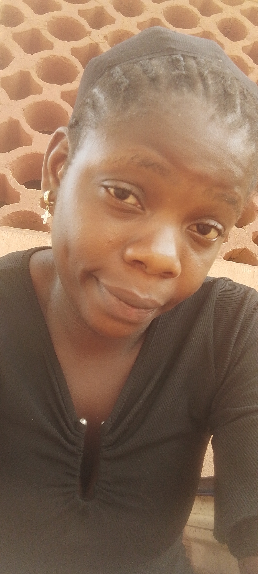 A medium shot depicts a young woman with dark skin, looking directly at the camera with a slight smile. She is wearing a close-fitting black top with small silver embellishments at the neck. Her hair is dark and appears to be closely cropped or covered with a cap. A gold hoop earring with a cross charm dangles from her left ear. The background is a textured, reddish-brown wall composed of hollow, cylindrical bricks. The location is identified as Unknown, Unknown.