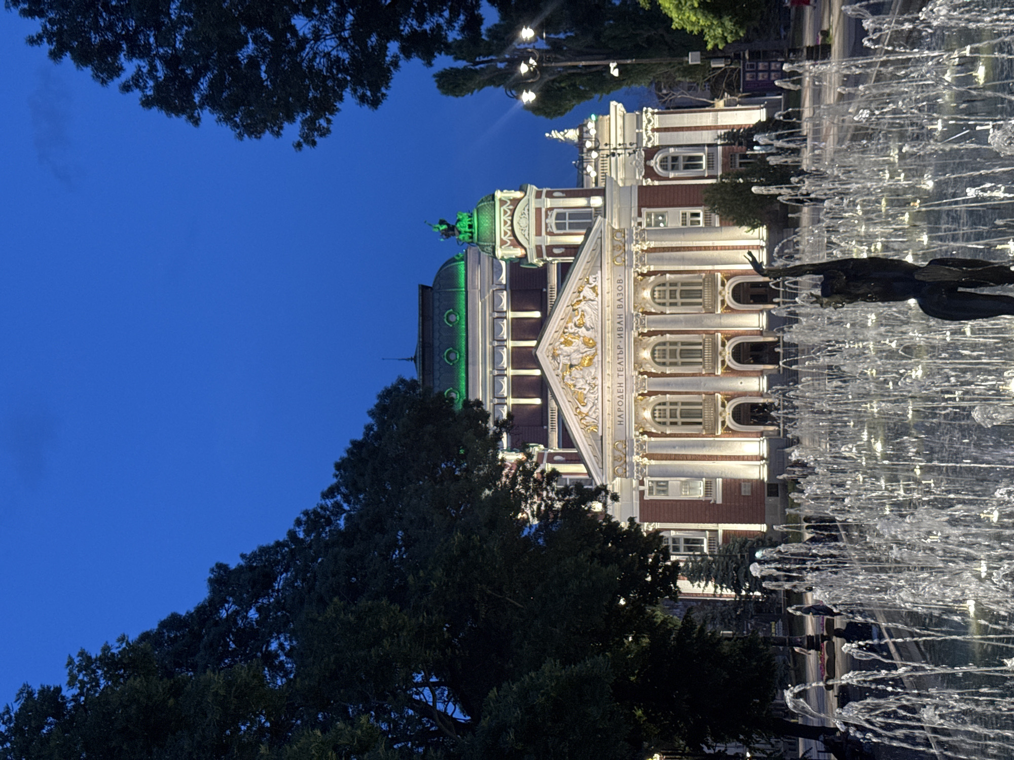 The image shows the Ivan Vazov National Theatre in Sofia, Bulgaria, at dusk. The theatre is a grand neoclassical building with a red brick facade, white columns, and ornate gold detailing. It is illuminated by warm lights, and the green-domed roofs are visible against the deep blue sky. In the foreground, a fountain is in operation, with numerous jets of water creating a dynamic display. A dark bronze sculpture of a dancer stands in the center of the fountain, its pose graceful and expressive. Several people are seated on benches in the background, enjoying the evening. The scene is framed by lush green trees on either side, adding to the serene atmosphere.