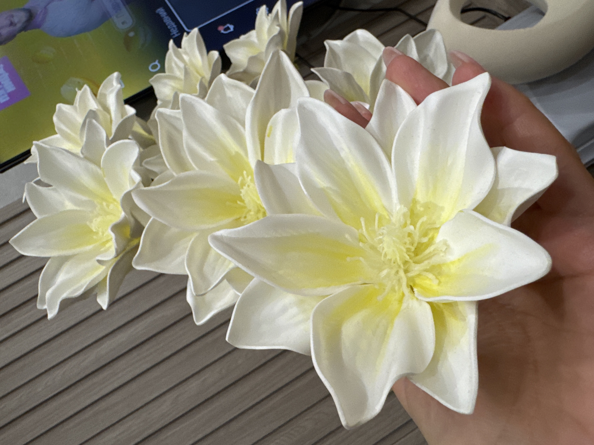 In this close-up, high-angle shot, a person's hand with light skin and natural-colored, long nails gently holds a cluster of artificial white and yellow flowers. The flowers, with their delicate, layered petals, are arranged in a diagonal line, with the closest one prominently featured in the foreground. The background consists of a textured wooden wall with vertical slats and a television screen displaying a person and text in a foreign language. The overall scene is well-lit, highlighting the intricate details of the flowers and the texture of the background elements.