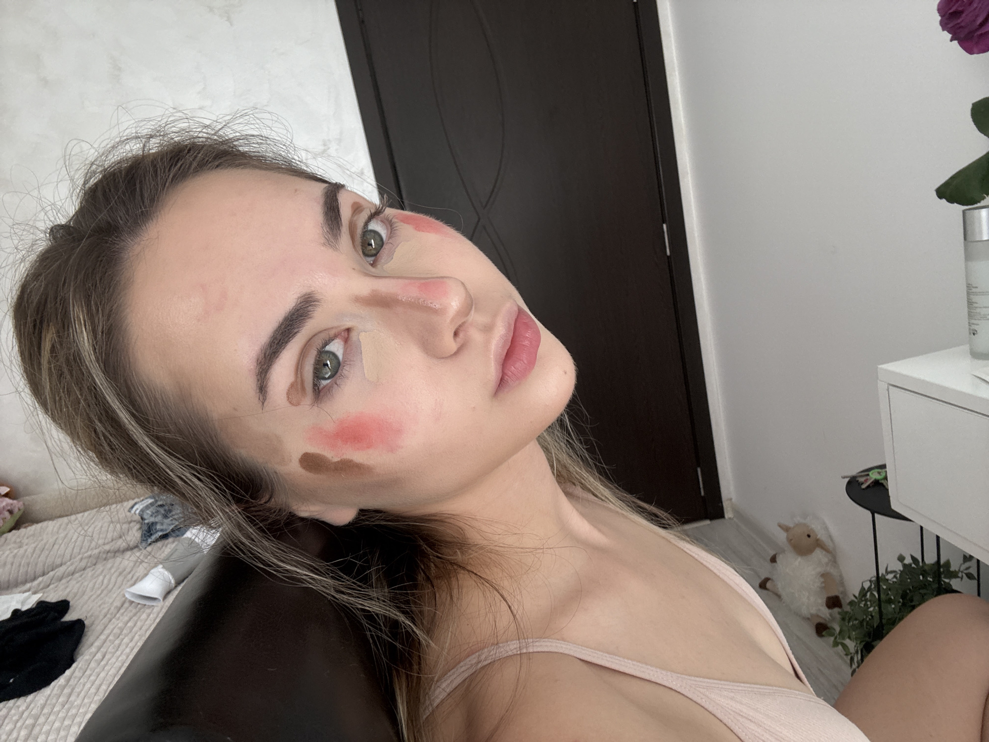 A young woman is positioned in the foreground, her face angled towards the camera. She appears to be in the process of applying makeup. Patches of different colored cosmetics are visible on her face, including concealer under her eyes and on her nose, as well as blush and what appears to be contour on her cheeks. She is wearing a light pink tank top.
Behind her, a dark brown door is visible, contrasting with the light-colored wall. A bed with rumpled covers and some items on it is to the left of the woman. To the right, there is a white cabinet with a purple flower on top, and a small stuffed sheep on the floor.
The setting appears to be inside a bedroom. There is no evidence to indicate that this image was taken in Blagoevgrad, Bulgaria.