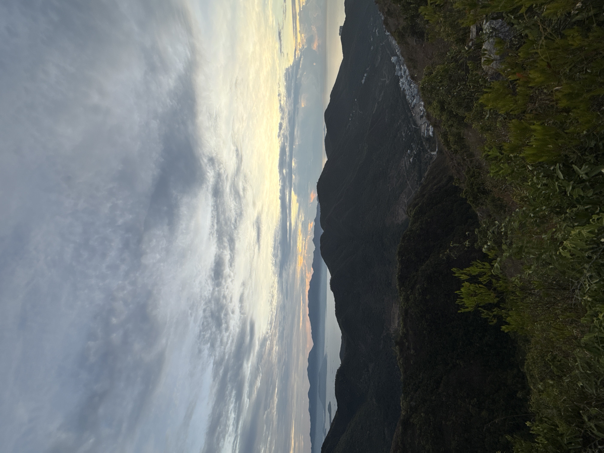 The image shows a view from a mountain overlooking the Bac Nha Trang Ward in Vietnam during a sunset. The sky is filled with layered clouds. The mountains are silhouetted against the sky. Vegetation is visible on the right side of the image. A small group of buildings can be seen on the mountainside.