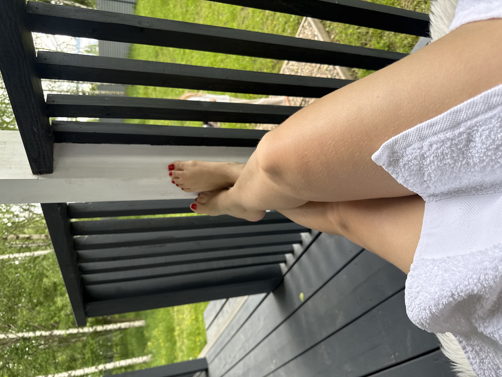 The image shows a pair of legs crossed and resting on a black wooden deck or porch. The person appears to be wearing a white towel or robe. The toes of the feet are painted red. Through the railing, grass and trees are visible in the background. Based on the location provided (Buzhaninovo, Russia), it is likely a rural or suburban setting with greenery.