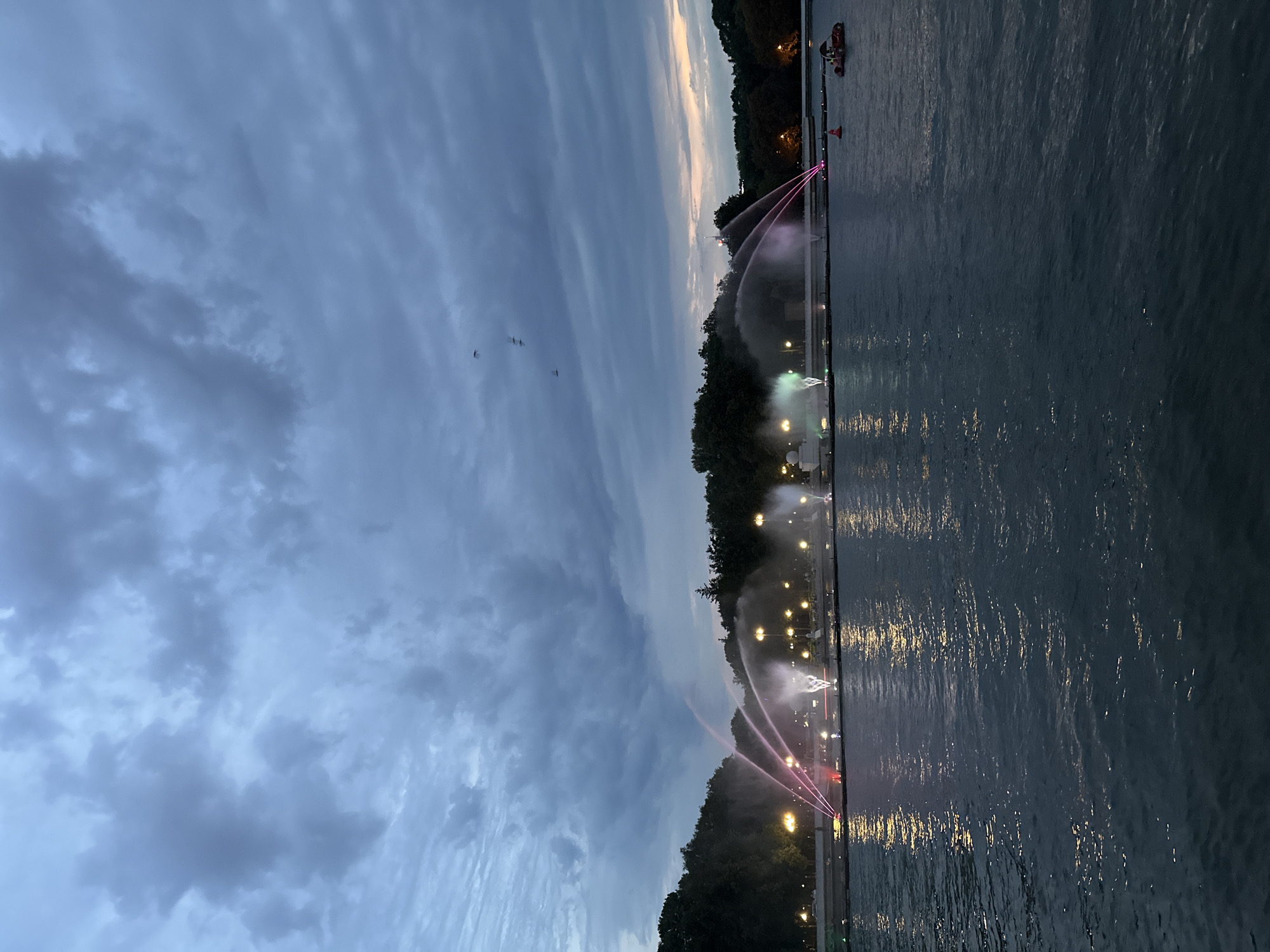 The image depicts a view across a body of water towards a fountain display and park area in Minsk, Belarus, likely during the evening or twilight. The sky is overcast with heavy, dark clouds. A line of illuminated fountains sprays water upwards, some with colored lights, creating a dynamic display. Behind the fountains, a tree-lined embankment is visible, dotted with streetlights. On the water, two small boats are present. One is a red and white buoy, and the other is a pedal boat with two occupants. The water itself is dark and reflects the lights from the embankment and the fountains.