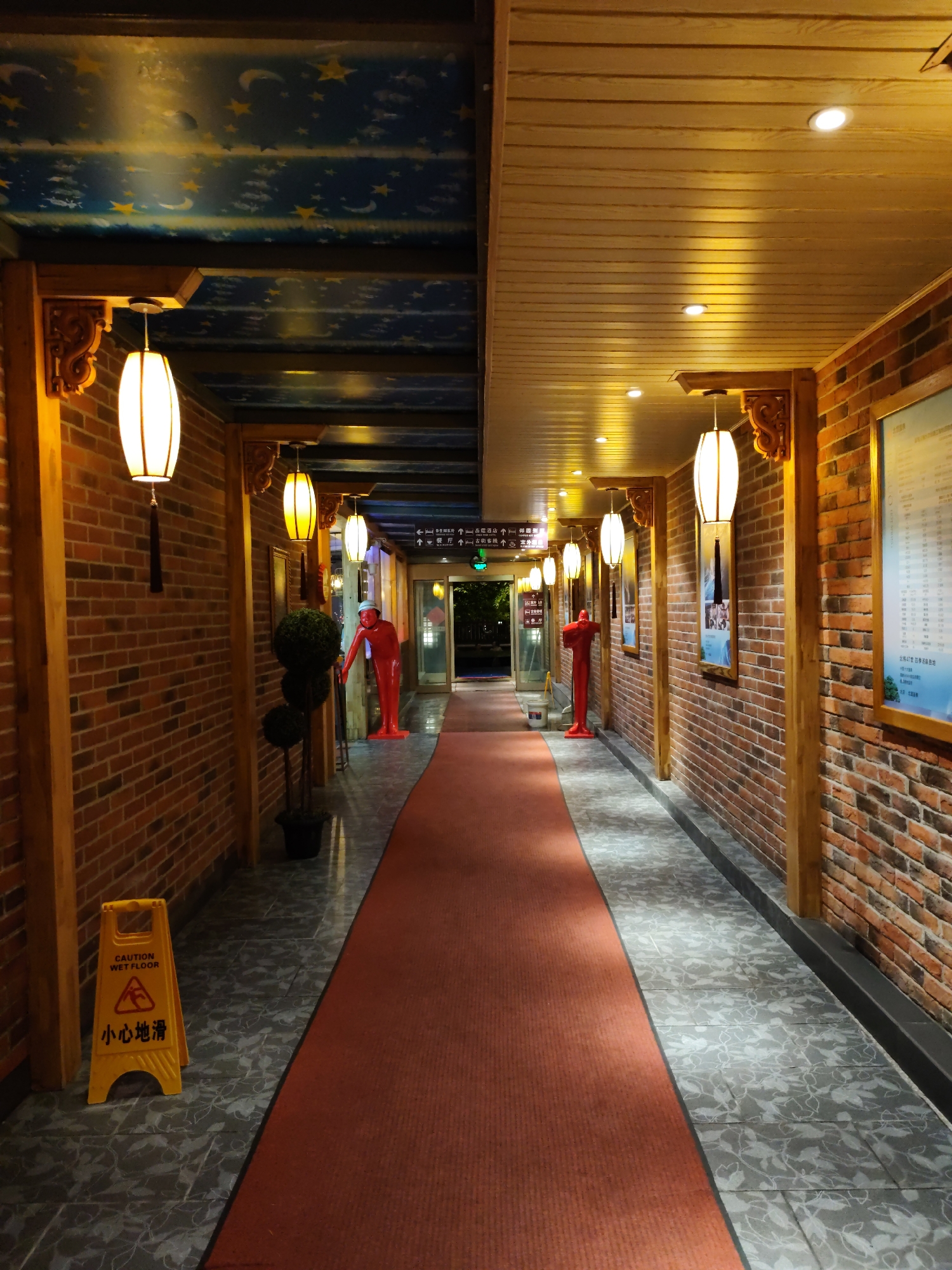 The image depicts a well-lit hallway with red brick walls and a wooden ceiling. A red carpet runs down the center of the hallway, flanked by grey patterned tiles. Several ornate lanterns hang from the ceiling, casting a warm glow. On the left side of the hallway, there is a yellow "Caution Wet Floor" sign with Chinese characters. Red, human-shaped statues are placed at intervals along the hallway. The ceiling on the left side is painted with a night sky motif featuring stars and crescent moons, while the ceiling on the right is made of horizontal wooden planks with recessed lighting. A framed informational poster is mounted on the brick wall on the right side. The hallway appears to lead towards a more open area with glass doors visible in the distance. The overall impression is one of an interior walkway, possibly in a hotel, entertainment venue, or a public building in 林甸镇, China.