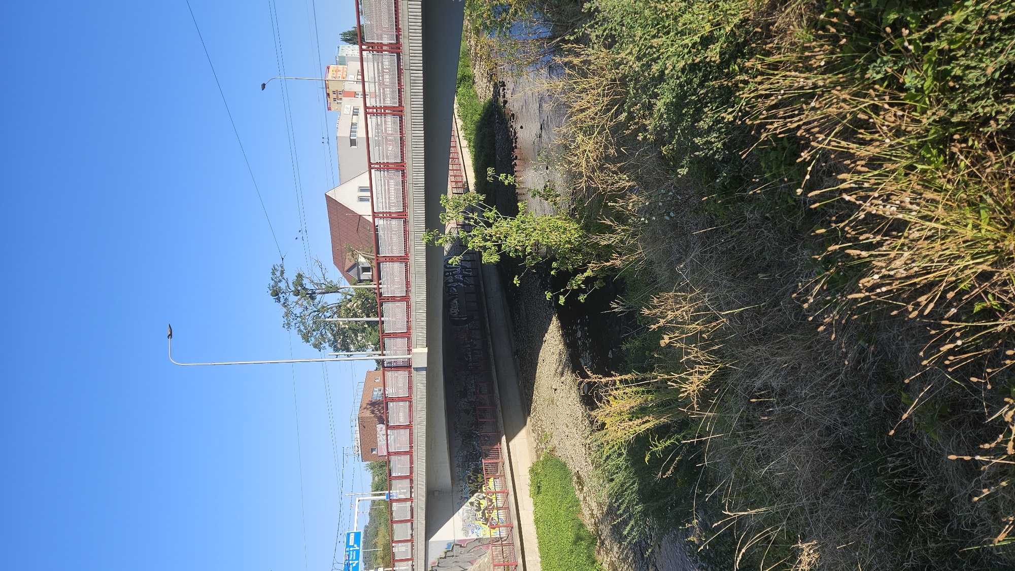 The image shows a sunny day in Prešov, Slovakia, with a concrete bridge spanning a dry, rocky riverbed. The bridge is adorned with red railings and graffiti on its underside. On the left side of the image, a blue road sign is visible. Buildings with red roofs and modern structures are situated behind the bridge, alongside trees. A streetlamp stands tall against a clear blue sky. The foreground is filled with dry grass and some green bushes, suggesting a natural area within the urban landscape.