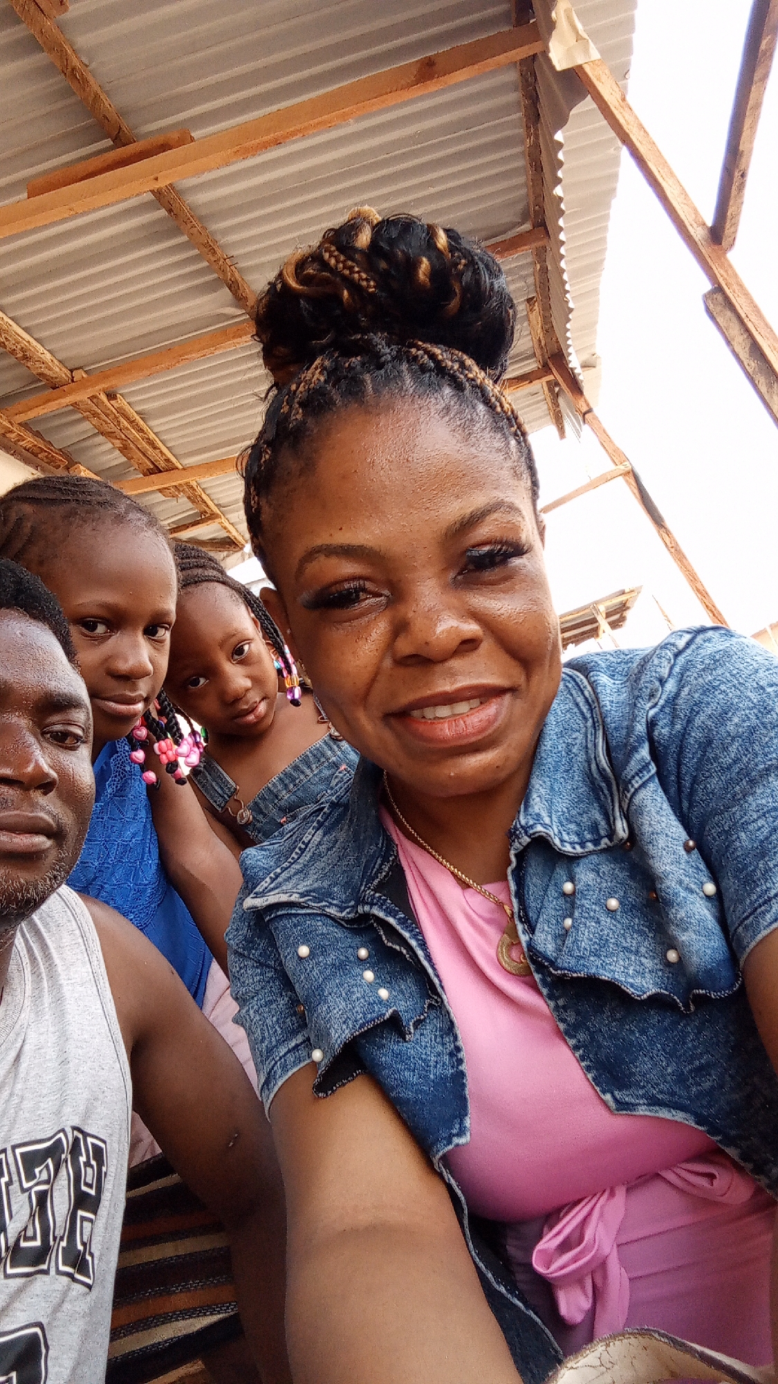 The image captures a selfie featuring a group of people in Sabon Tasha, Nigeria. A woman with braided hair styled in a high bun and wearing a pink top with a denim jacket embellished with pearls is in the foreground. Behind her, two young girls are visible, one in a blue top and the other in a denim outfit with colorful beads in her hair. A man in a grey tank top is partially visible on the left side of the frame. The backdrop consists of a corrugated metal roof supported by wooden beams, indicating an outdoor or semi-outdoor setting. The lighting suggests it is daytime.