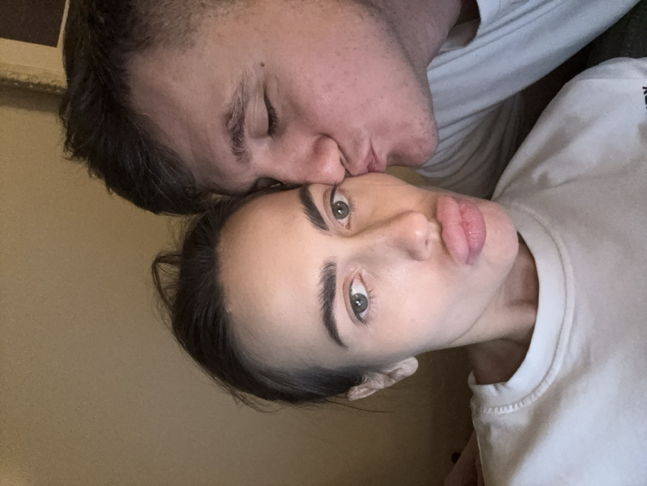 A close-up, head-and-shoulder portrait features two individuals, a female and a male, in an indoor setting within the city of Venice, Italy. The female, positioned on the left, has dark hair pulled back, distinct eyebrows, green eyes, and her lips are puckered into a kissing pose as she looks directly at the camera. She wears a light-colored, possibly white, crew-neck sweatshirt. The male, positioned on the right, has short dark hair and his eyes are closed. He is shown in profile, leaning towards the female, and is kissing her right cheek. He wears a light-colored top. The background is a plain, light-brown wall, suggesting an intimate or private environment.