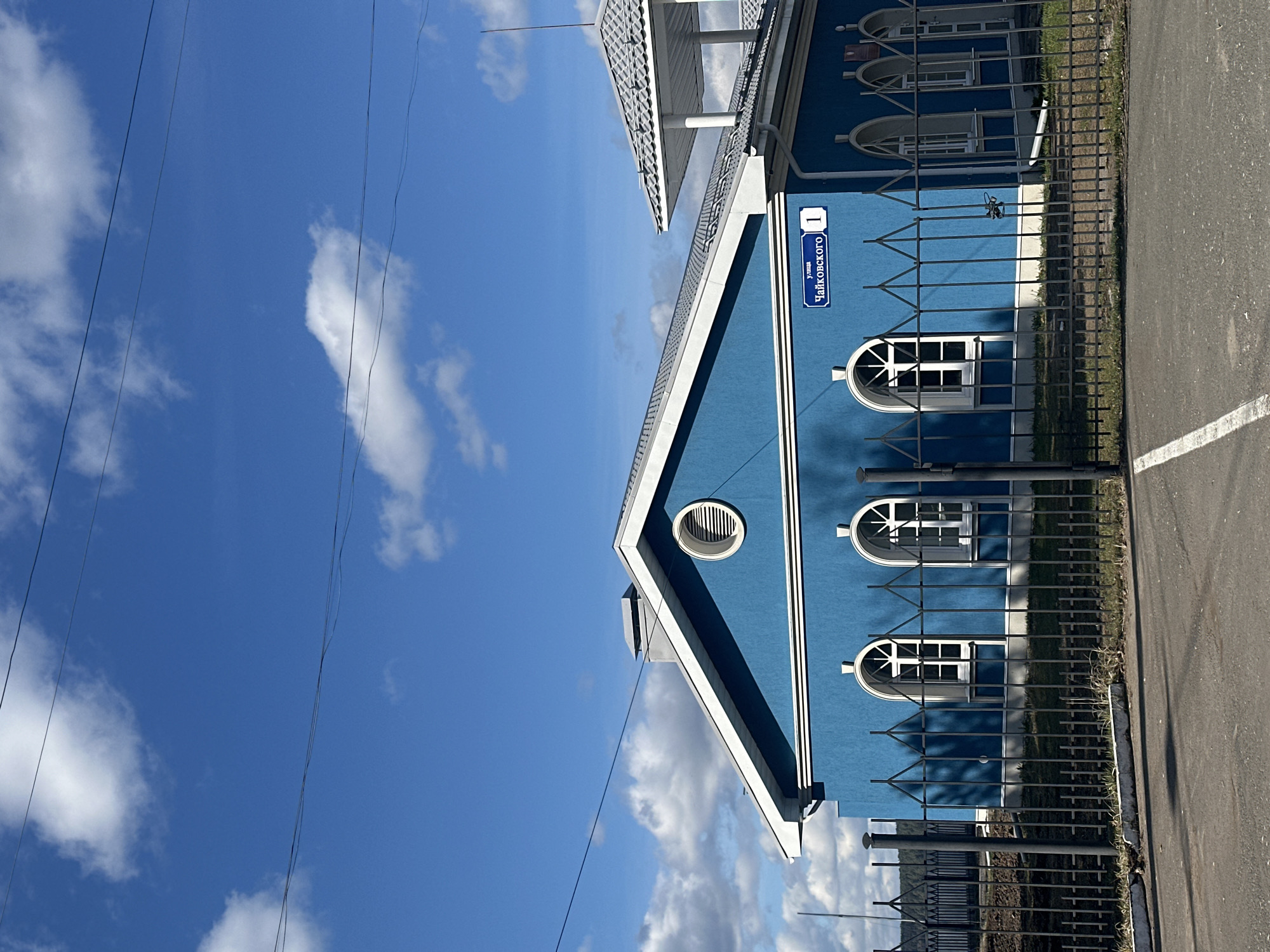 A low-angle, rotated view shows a light blue building against a partly cloudy sky. The building, located at 1 Tchaikovsky Street in Votkinsk, Russia, has a white roofline, a sign indicating its address, and several arched windows with white trim. A black wrought-iron fence runs along the building's perimeter next to an asphalt road. Power lines are visible against the sky.