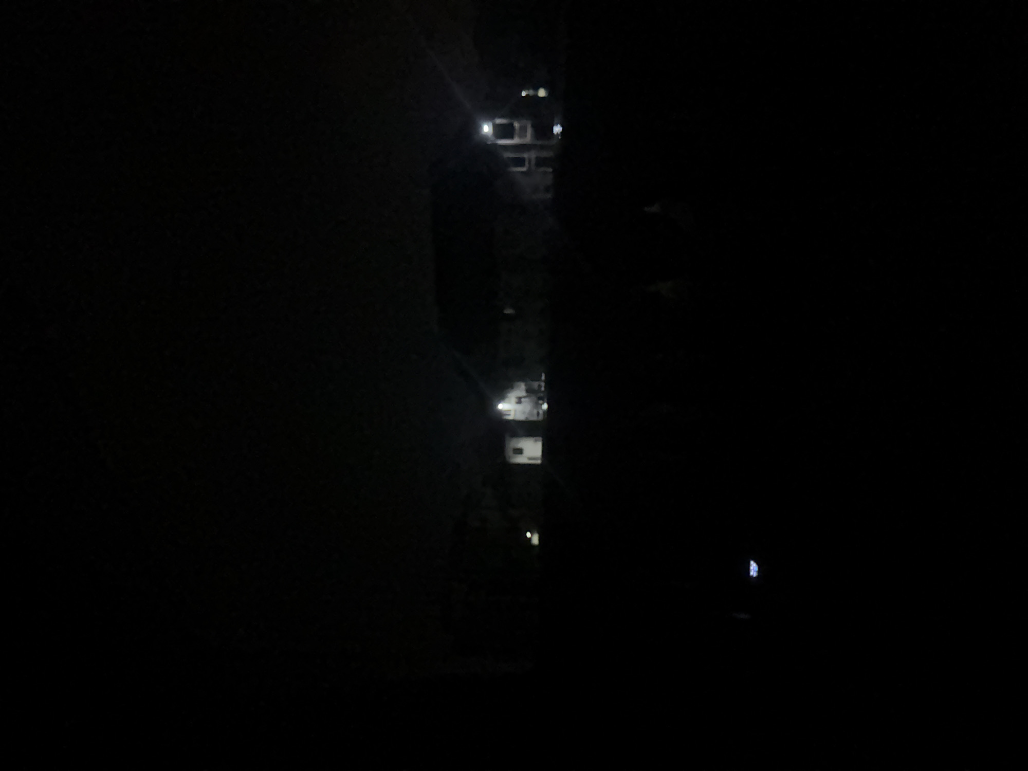 The image shows a vertical view of buildings at night in Ihiagwa, Nigeria. The structures are interspersed with foliage. The lighting conditions are low, with a few visible lights within the buildings.