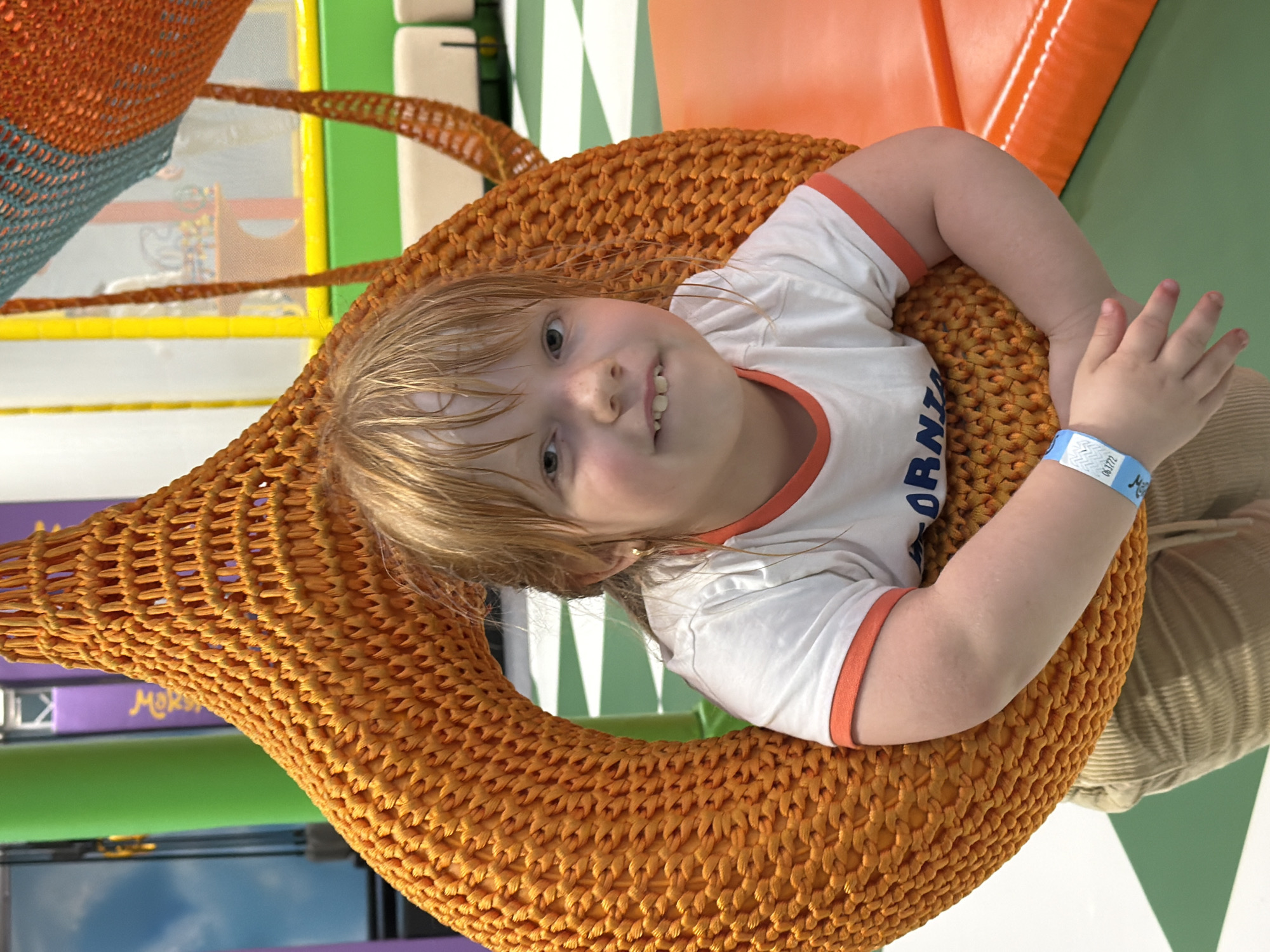A young girl with blonde hair is sitting in a large, orange, woven pod-like swing. She is wearing a white t-shirt with orange trim and a blue wristband with text and numbers. The background appears to be an indoor play area with bright colors, including green and yellow structures and a checkered floor. There are padded orange surfaces visible in the foreground. The image was taken in Tallinn, Estonia.