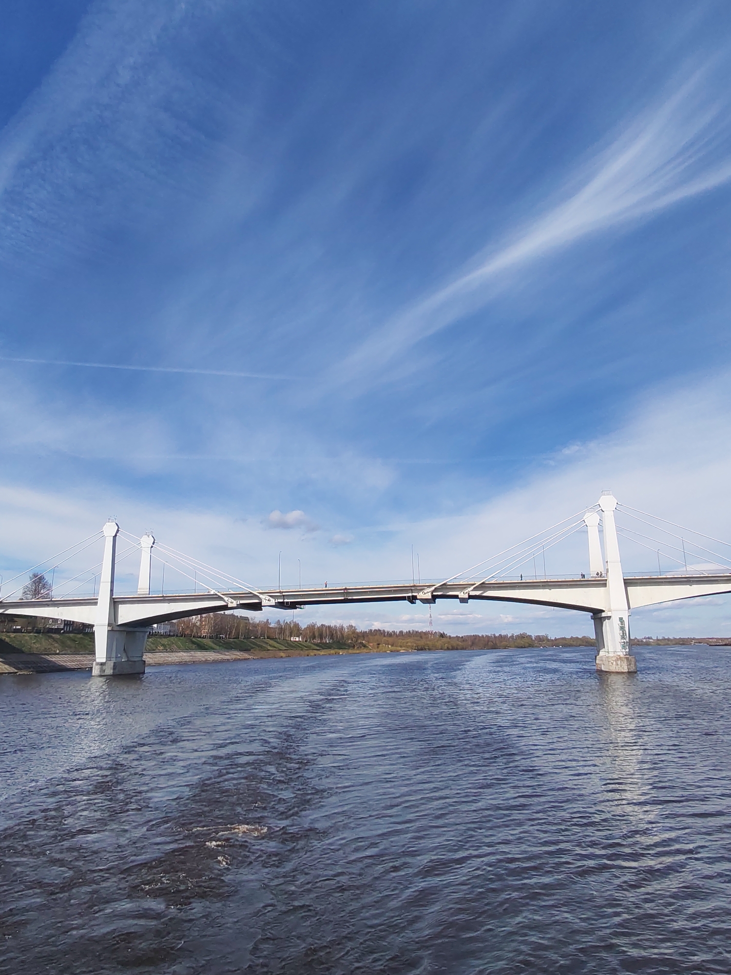 The image shows a cable-stayed bridge over a river in Kimry, Russia. The bridge is painted white and has two towers with a hexagonal shape at the top. Cables extend from the towers to support the bridge deck. There are people walking on the bridge. The river is dark with ripples. The far shore is visible with some trees and a low-rise cityscape. The sky is blue with wispy cirrus clouds.